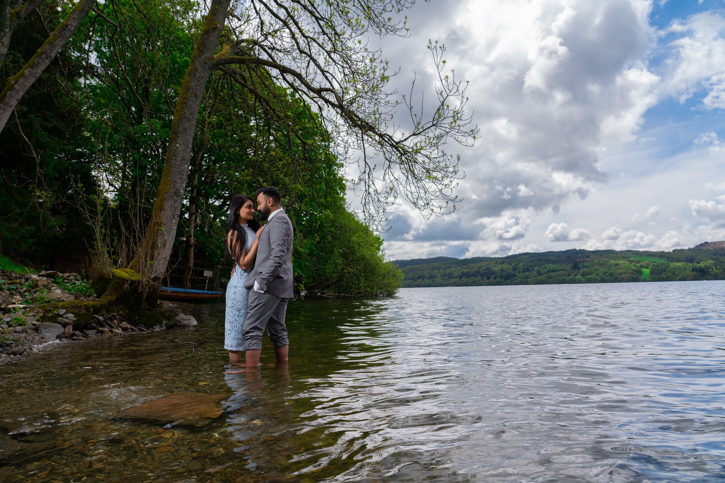 A couple stands close together in shallow water by a lakeside, with trees and hills in the background. They are looking into each other's eyes, dressed in formal attire; the woman in a lace dress and the man in a gray suit, with cloudy skies overhead.