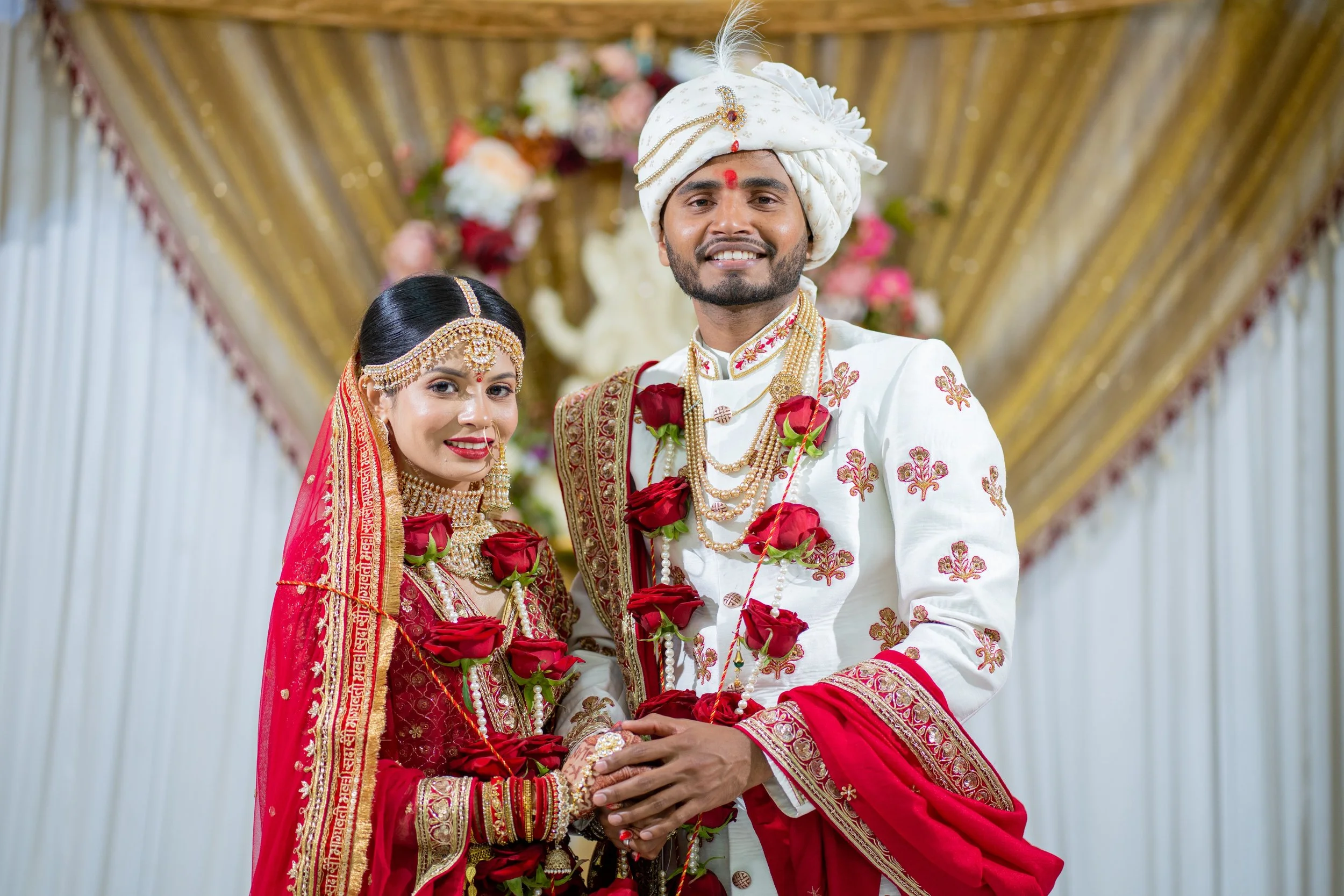 A smiling Indian bride and groom dressed in traditional wedding attire, standing together in front of a decorated background with floral arrangements and drapes.