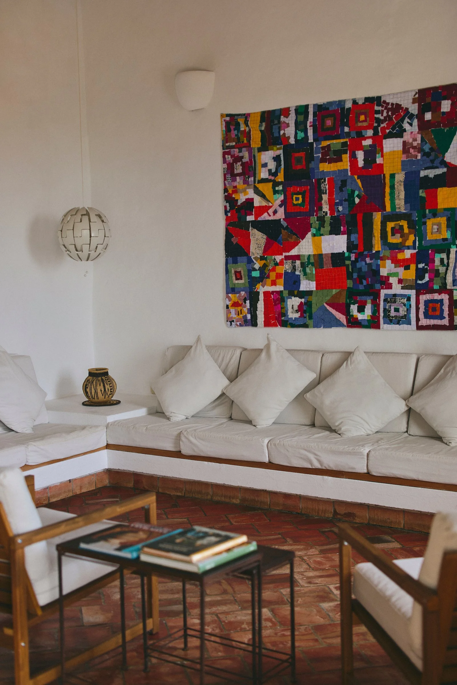 Shared living room at Casa Barichara with white seating, terracotta tile floors, books, regional decor, and a colorful textile artwork.