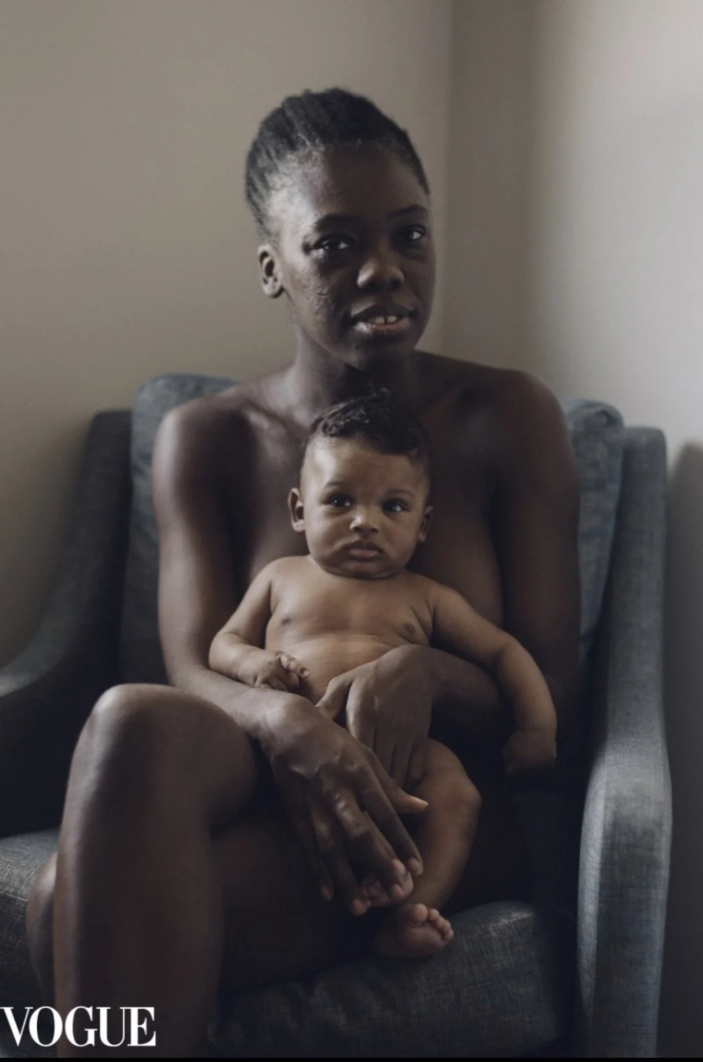 A woman sitting on a gray armchair holding a naked baby on her lap in a room with plain walls. The image has a VOGUE logo in the bottom left corner.