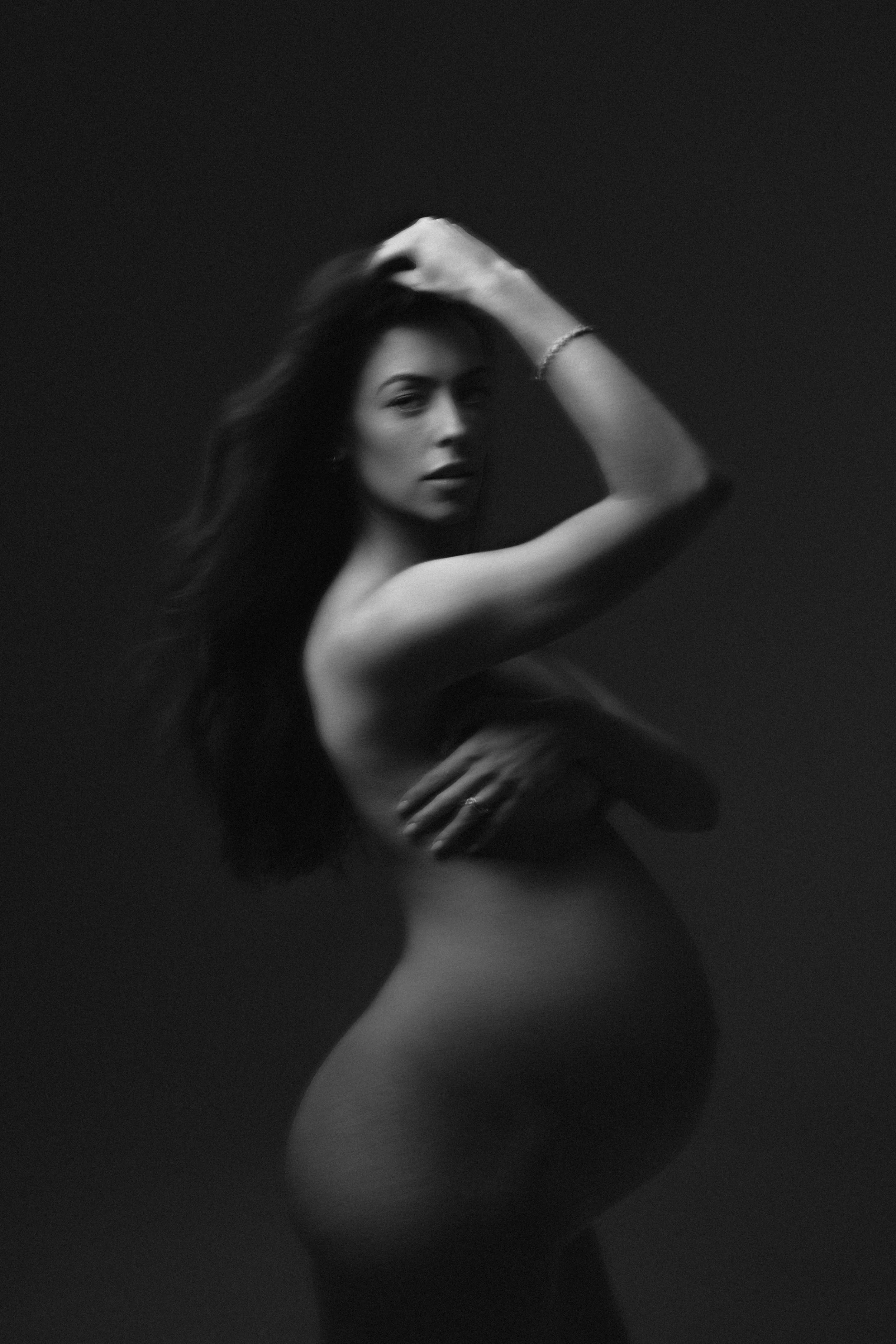 Black and white photo of expecting mother  with long hair, with one hand on her head and the other crossed over her chest.