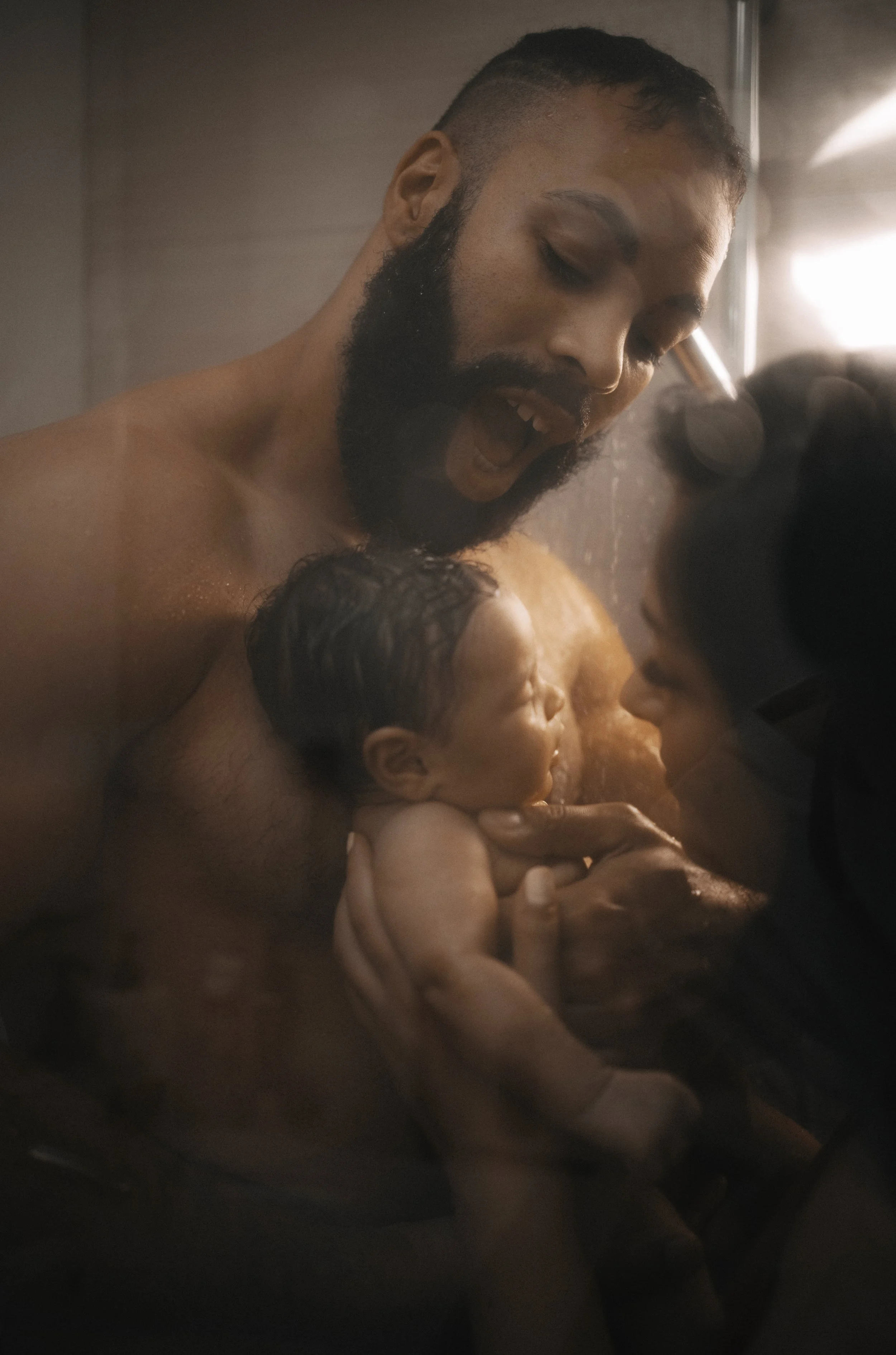A man with a beard and a woman hold and support a newborn baby in a shower, sharing a tender moment.