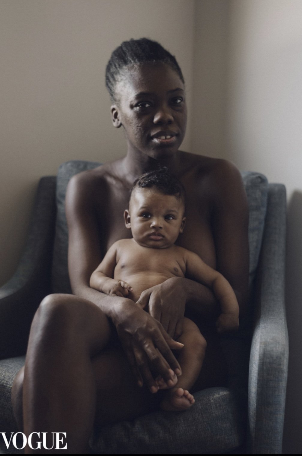 A woman holding a baby, both sitting on a gray armchair in a room with a neutral wall, with the Vogue logo in the bottom left corner.