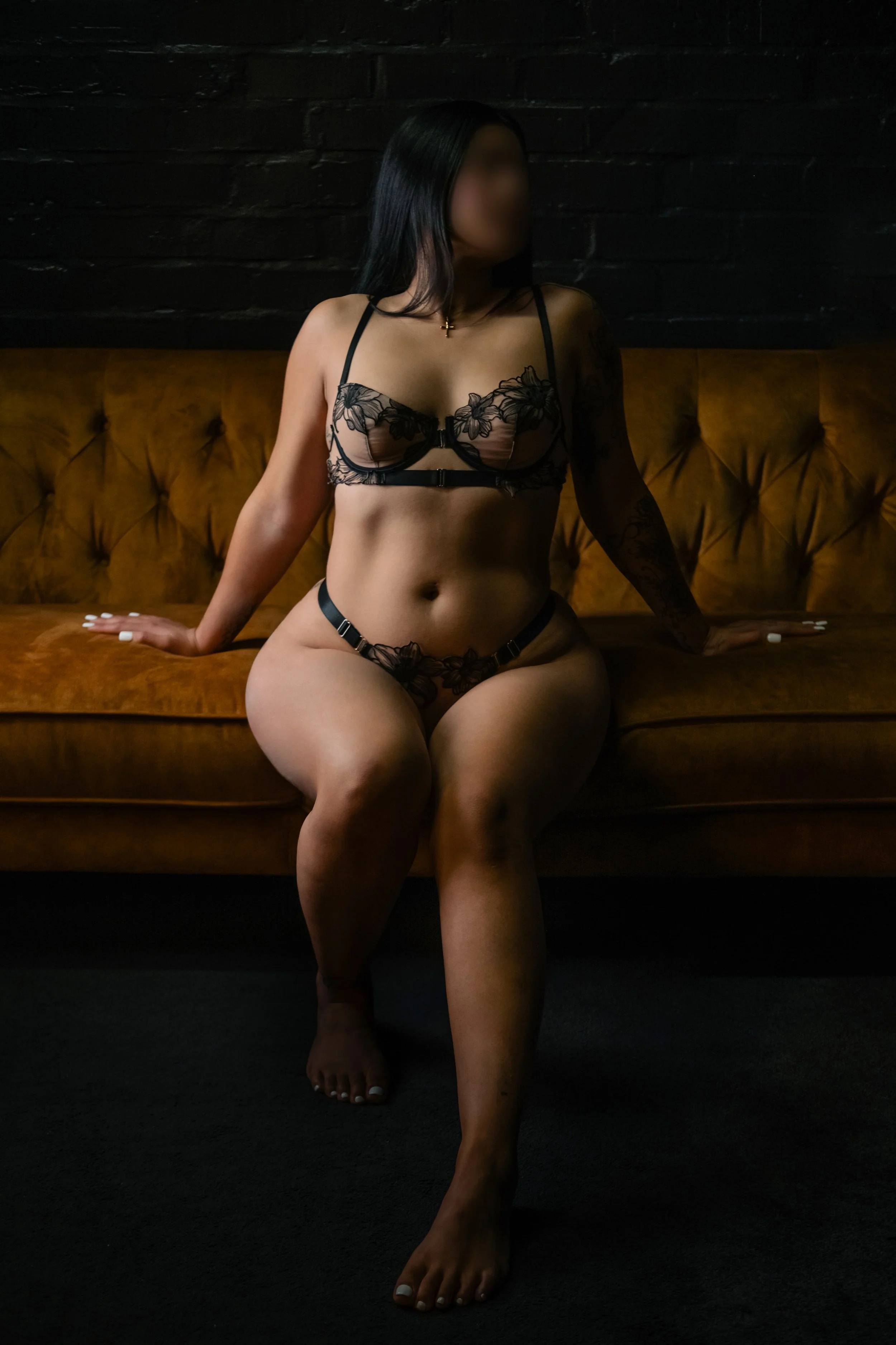 A woman wearing black floral lingerie sitting on a brown velvet couch against a black brick wall, with her face blurred, one hand on the couch, legs slightly apart, and toes painted white.