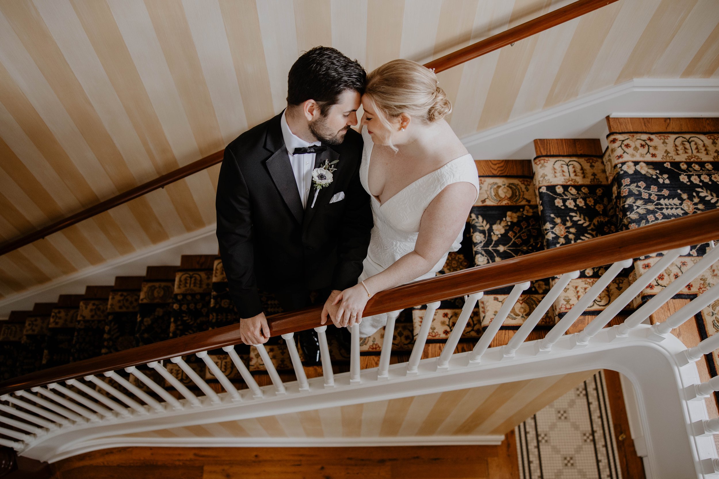 A bride and groom leaning foreheads together on a staircase landing, seen from above. The bride wears a white dress, and the groom wears a black tuxedo.
