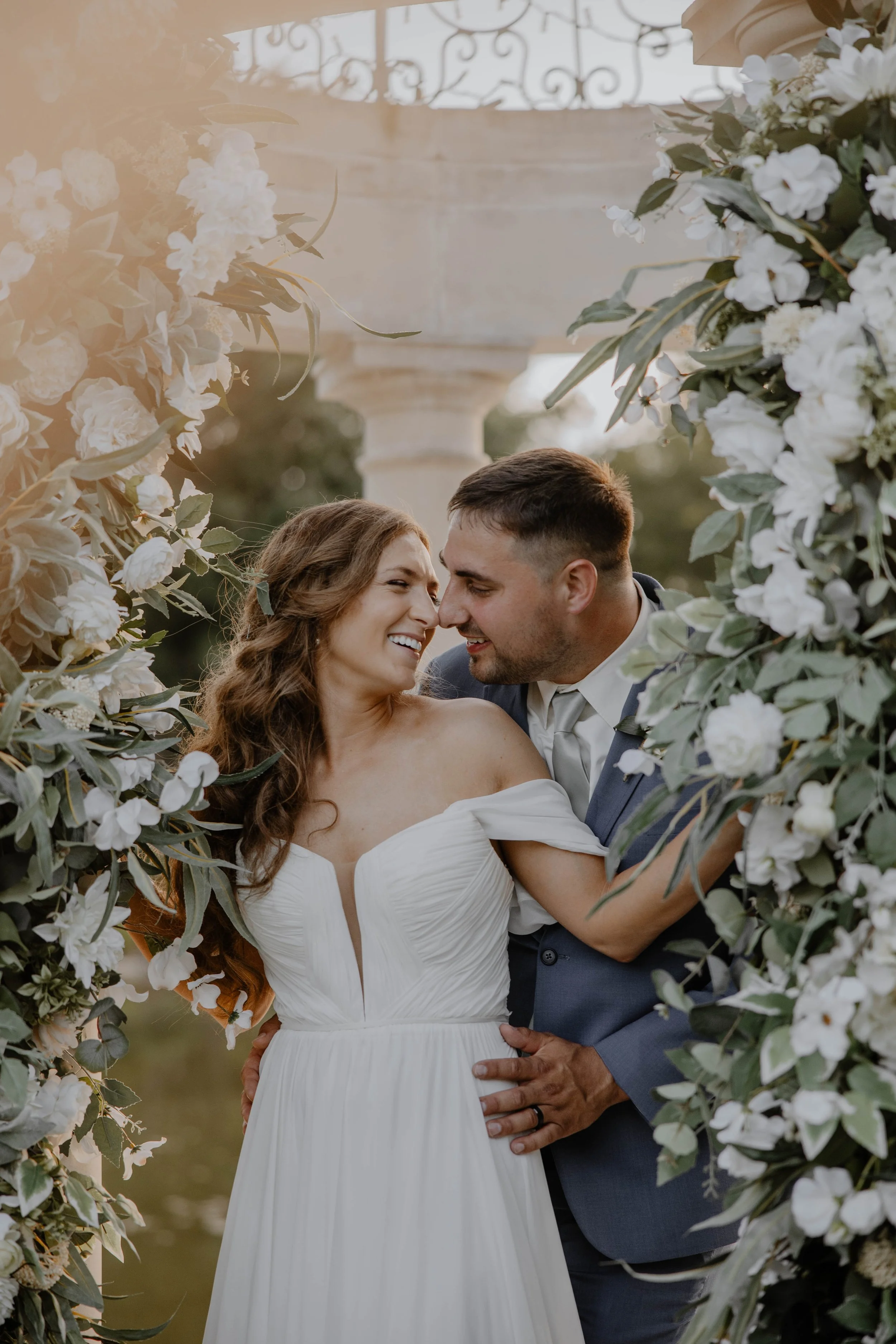 A newlywed couple in wedding attire sharing a joyful moment surrounded by white flowers and greenery during their outdoor wedding ceremony.