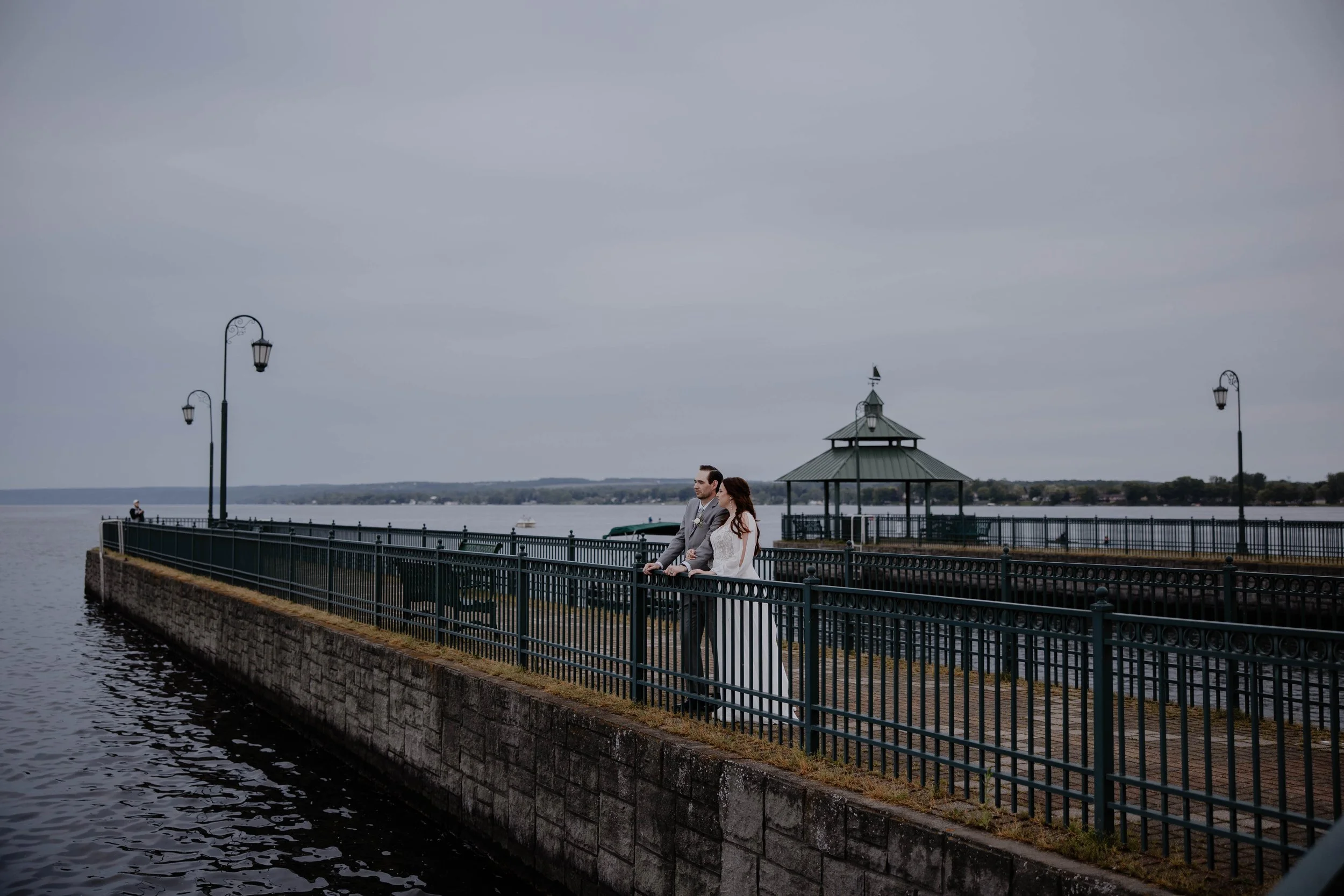 A couple in wedding attire standing on a waterfront pier during overcast weather, with a railing, lampposts, and a gazebo in the background.