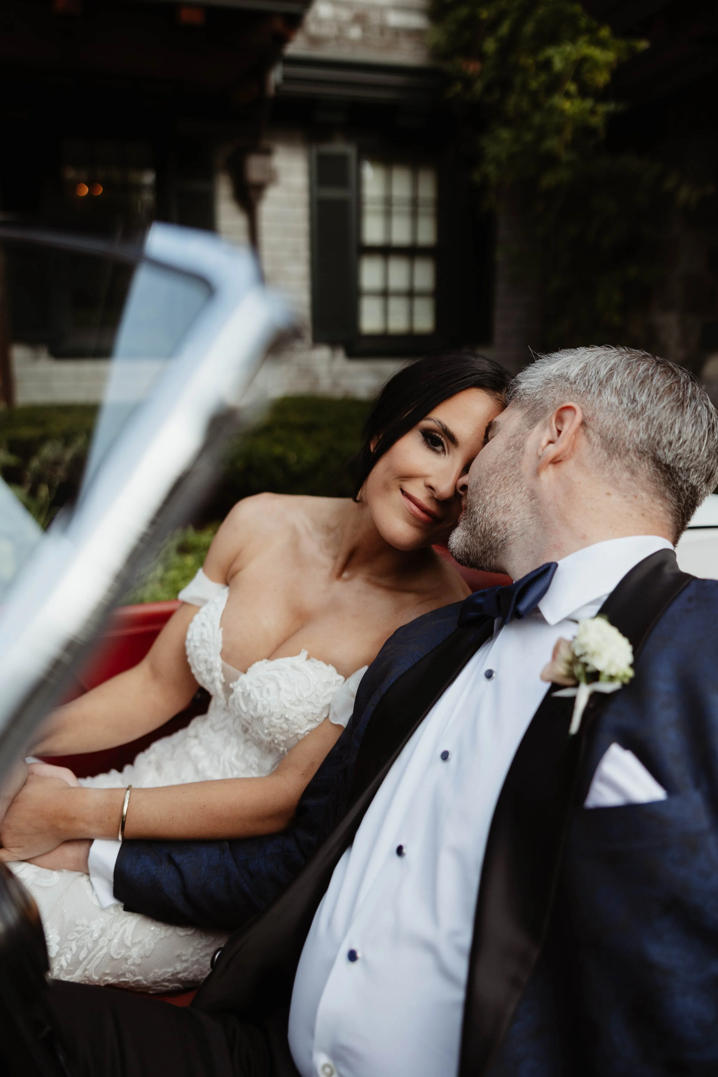A bride and groom sitting closely together in a vintage car, with the groom whispering to the bride who is smiling and leaning her head on his forehead. The bride is wearing a white wedding dress and the groom is in a tuxedo with a boutonniere.