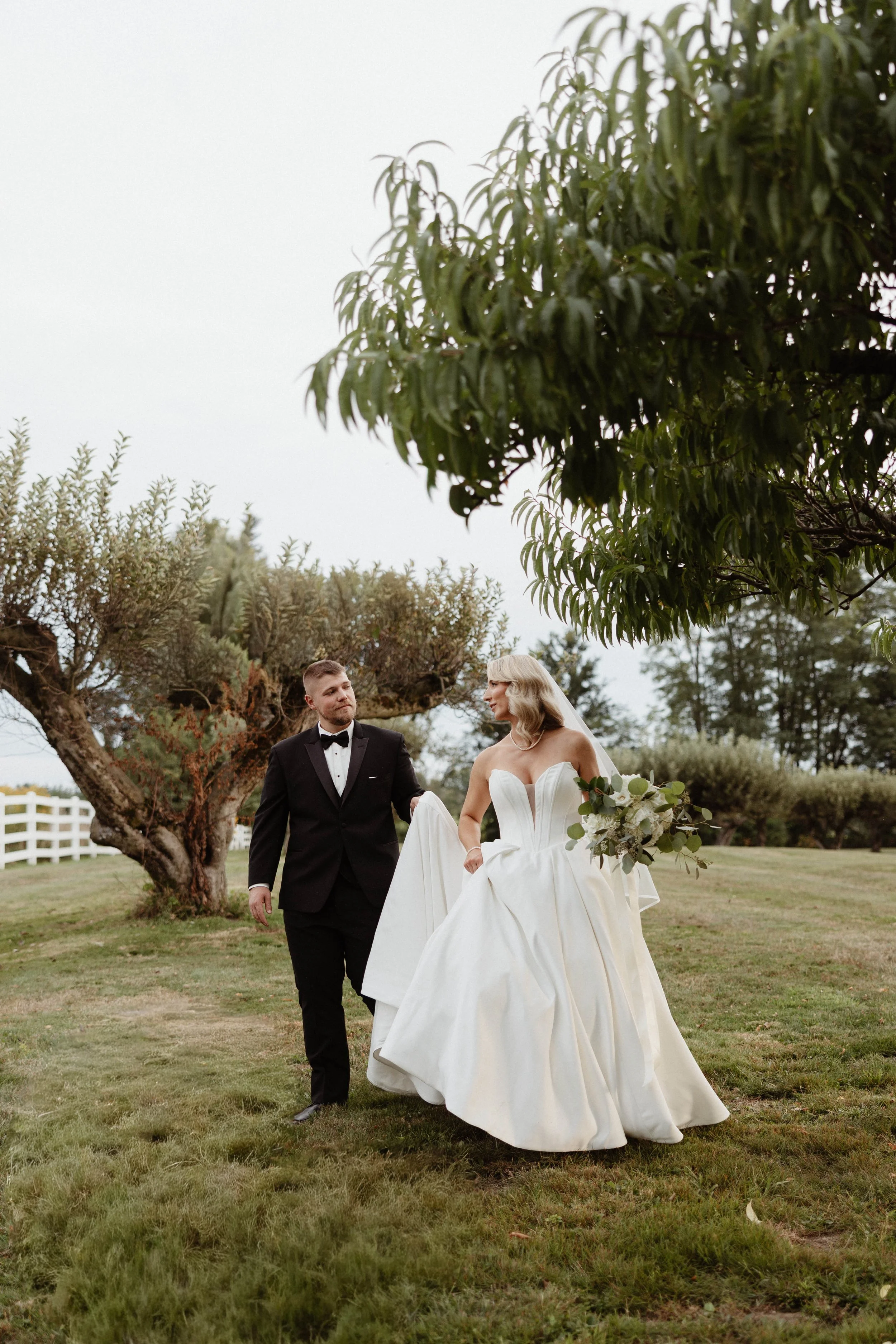 Bride and groom walking on grass outdoors, with bride in a white wedding dress holding a bouquet and groom in a black tuxedo, trees in the background.