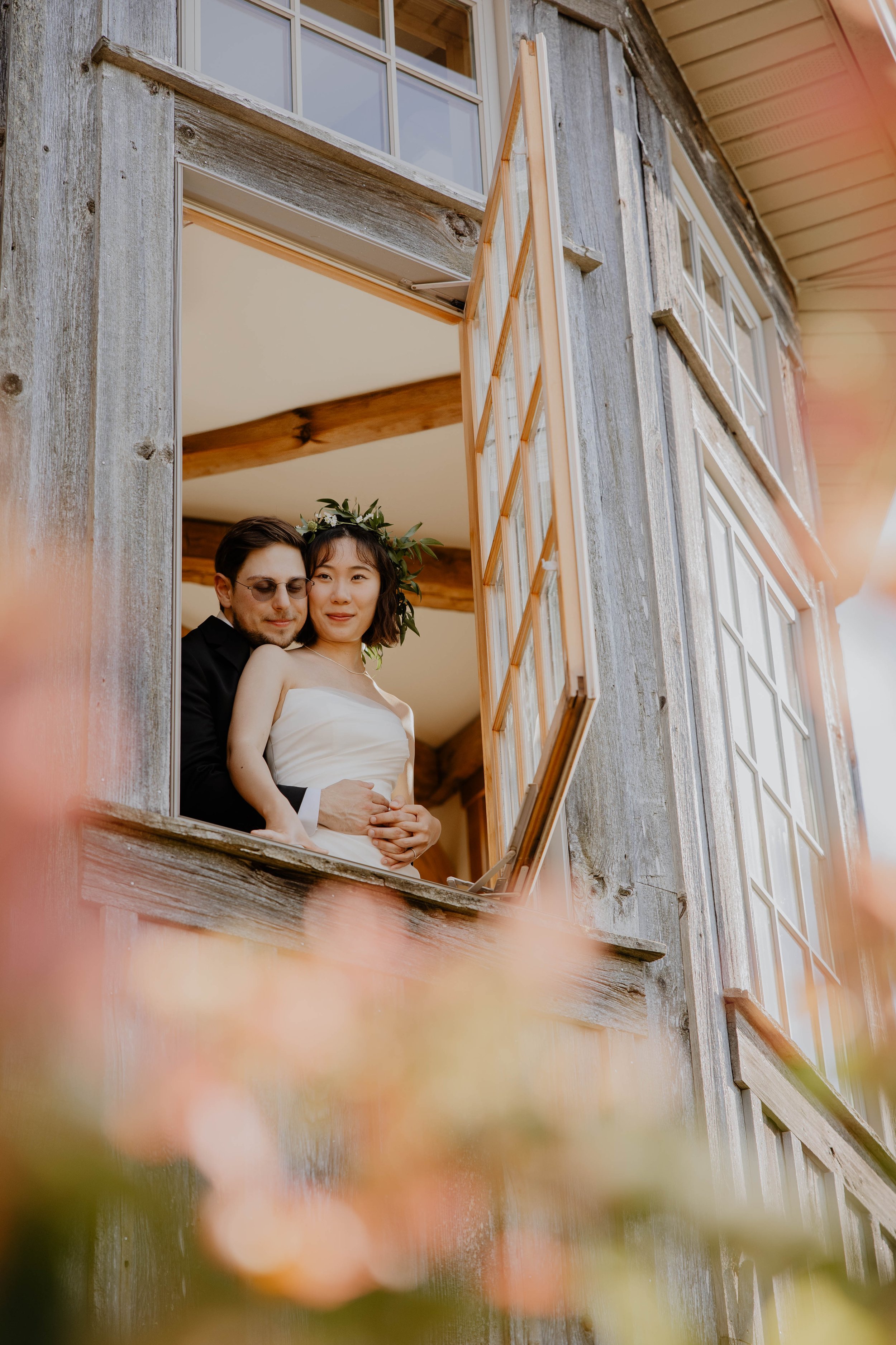 A couple, the bride wearing a white dress and a leafy headpiece, and the groom in a black suit, are standing together at a window of a rustic wooden building, looking out happily.