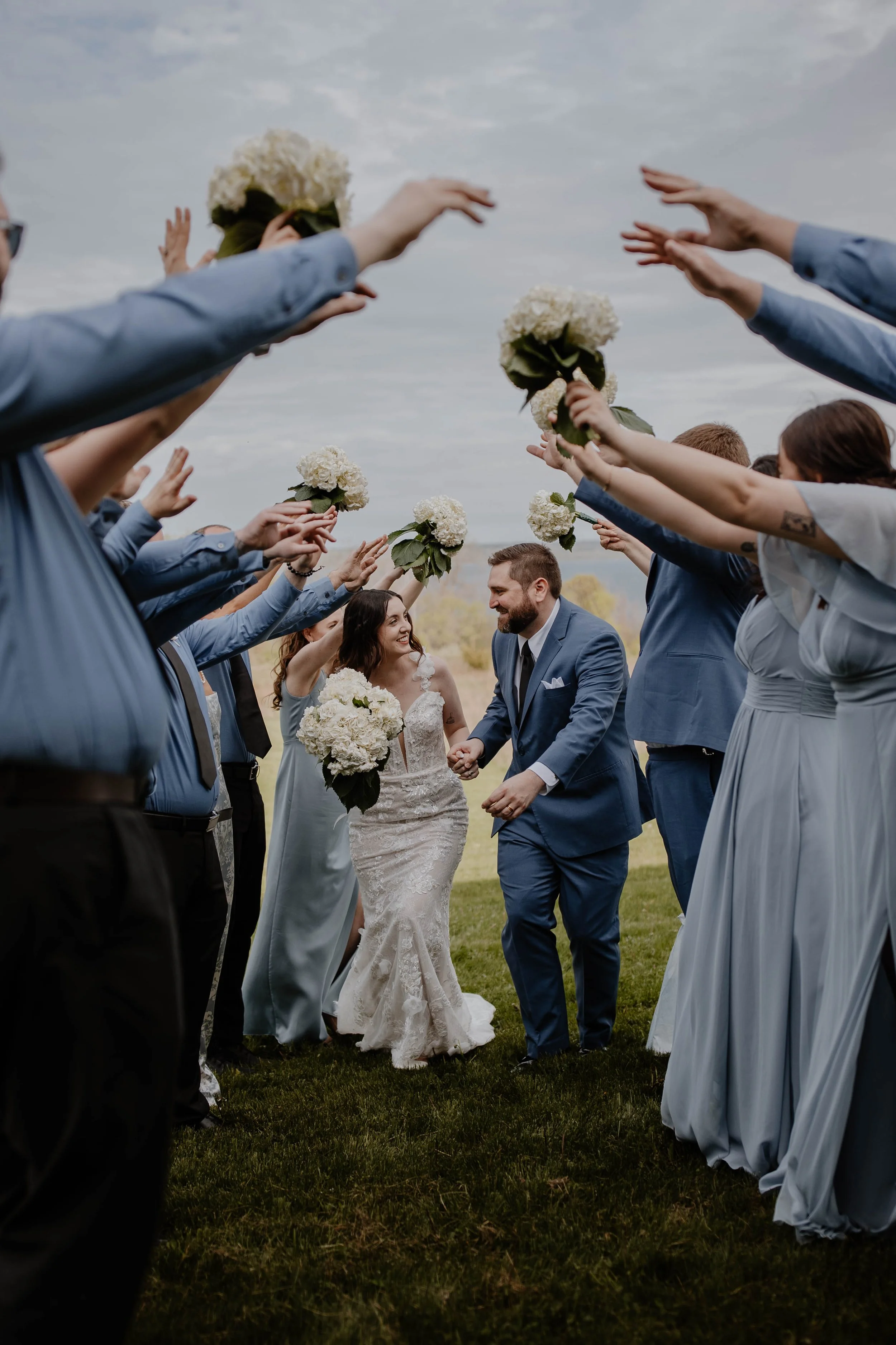 A bride and groom walking hand in hand through a tunnel of bridesmaids and groomsmen holding white flower bouquets and reaching towards them.