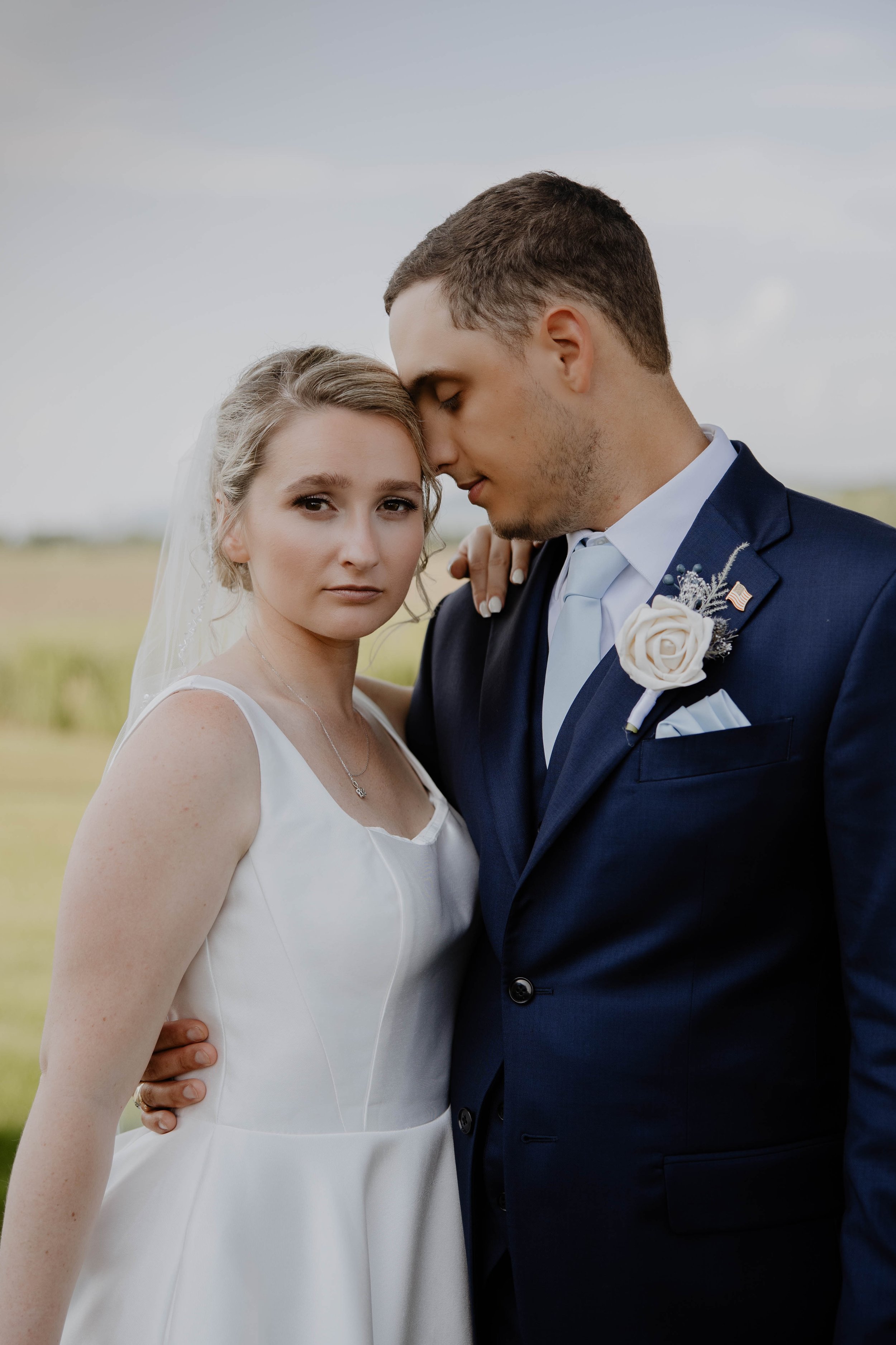 A bride and groom standing close together outdoors, with the groom leaning his forehead against the bride's temple. The bride is wearing a white wedding dress and a veil, while the groom is in a navy suit with a white shirt and tie, and a white rose 