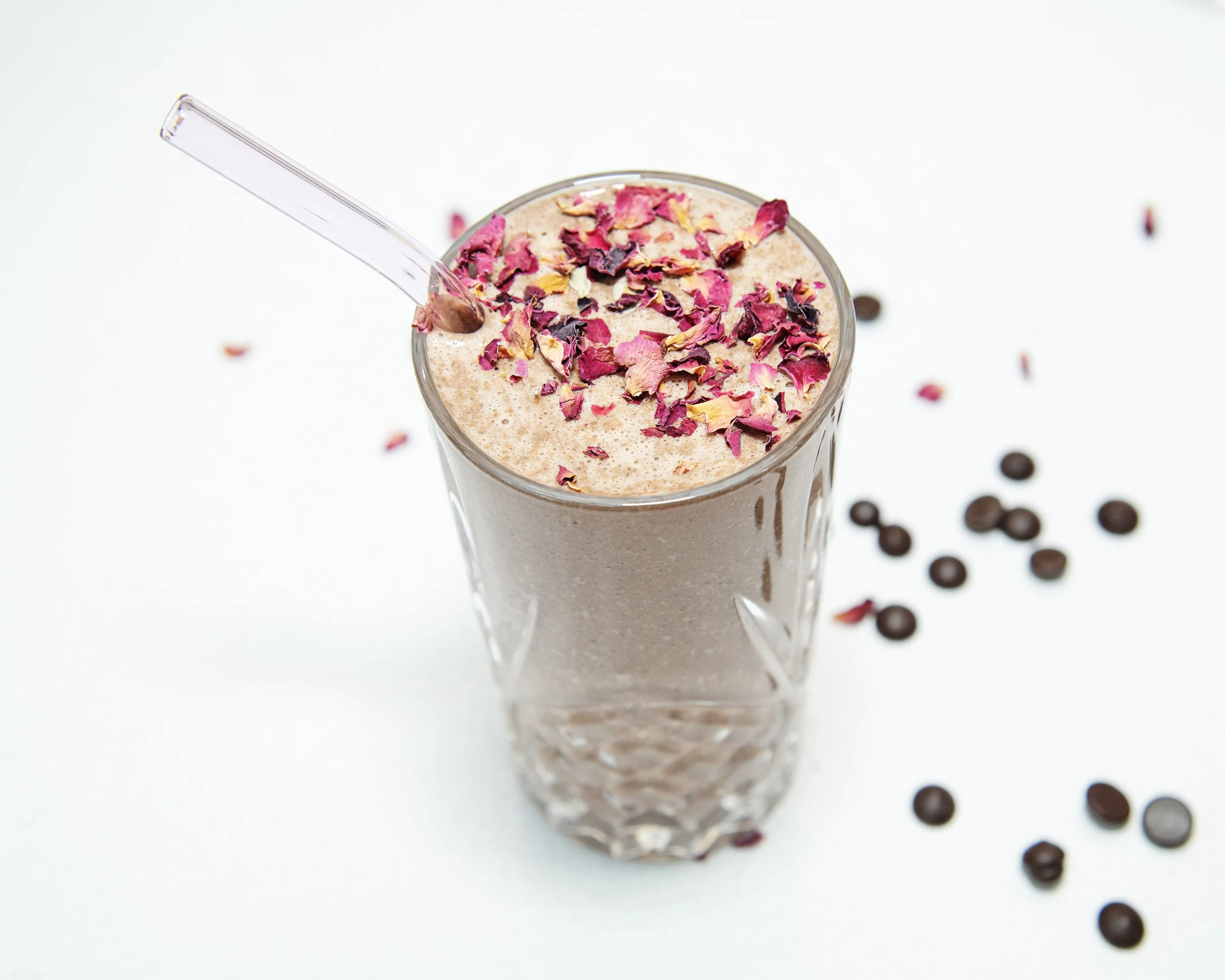 Glass of coffee smoothie topped with dried rose petals and a clear straw, surrounded by coffee beans on a white background.