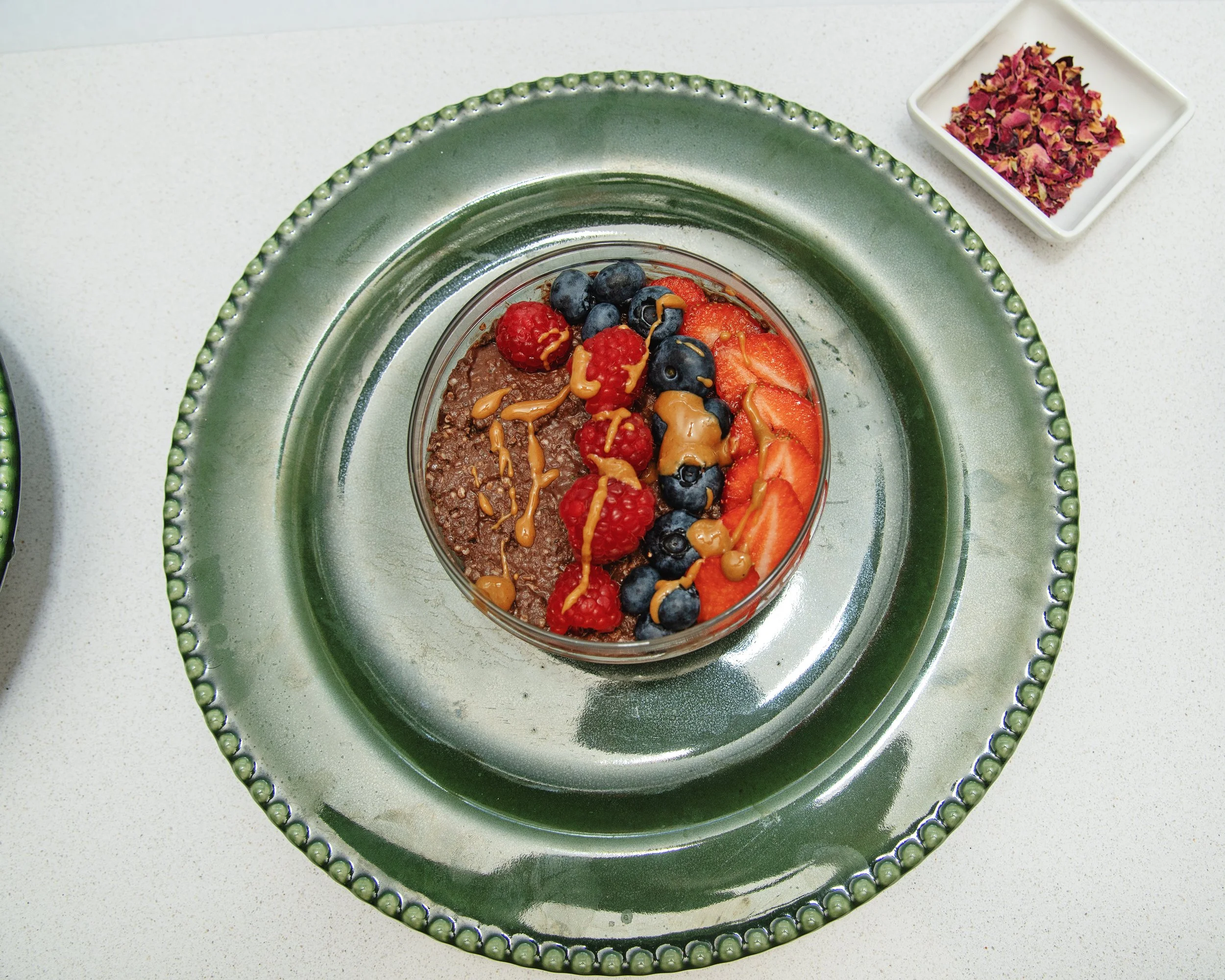 Bowl of chocolate chia pudding topped with strawberries, raspberries, blueberries, and peanut butter drizzle on green plate, next to rose petals in a white dish.