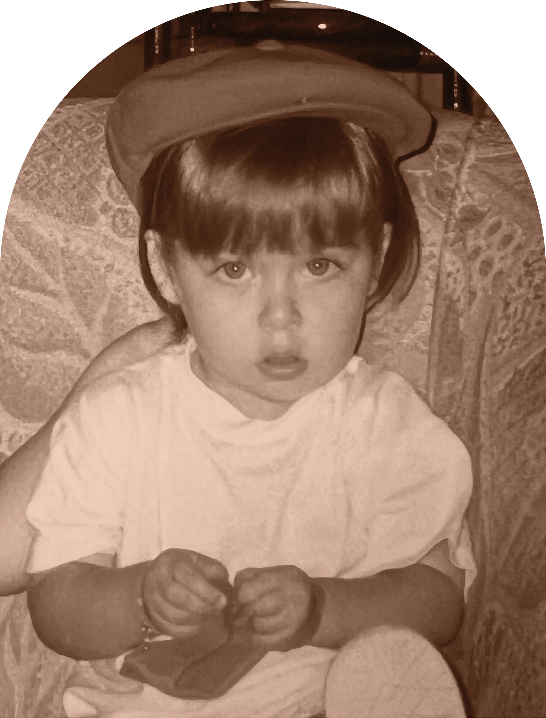A young child with short hair wearing a cap, sitting on a patterned couch, looking directly at the camera with a surprised expression and clenched fists.