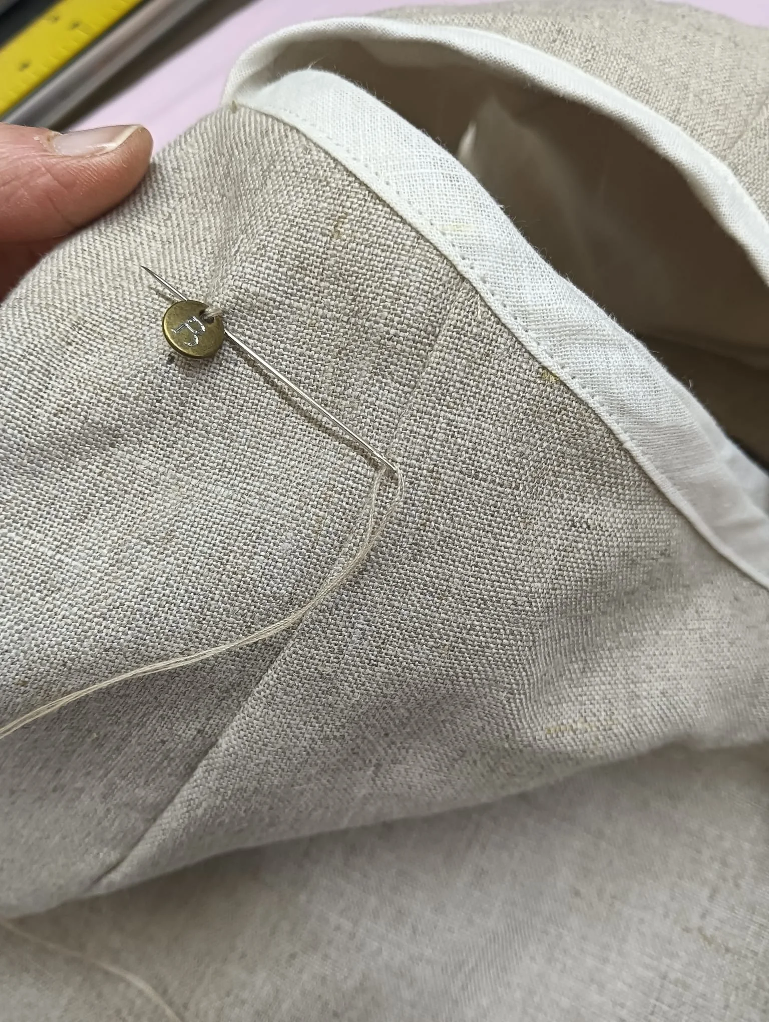Close-up of beige fabric with a pin and a small round metallic tag attached, showing a corner of a linen pocket or section.
