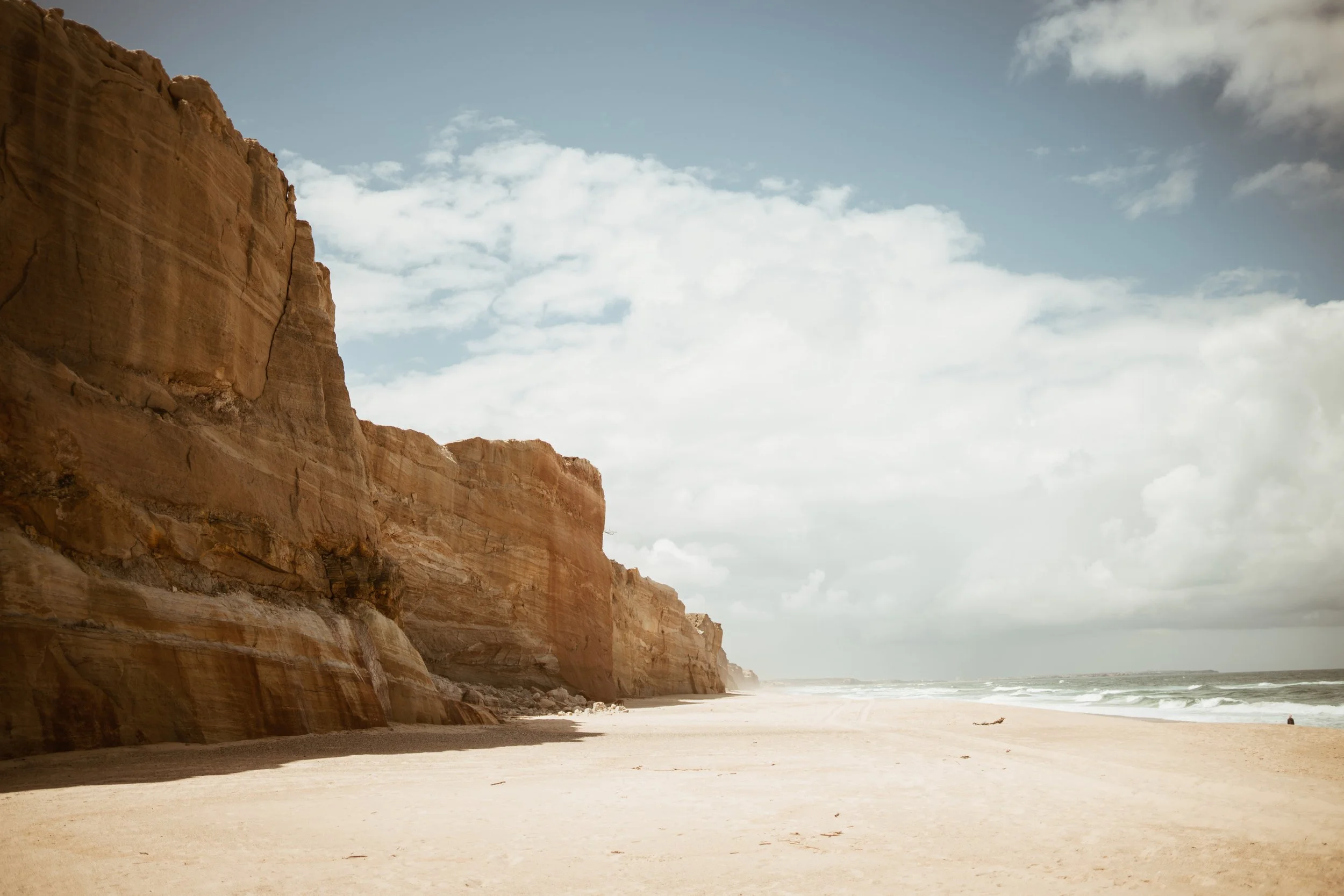 A beach with sandy shore, towering orange-brown cliffs on the left, cloudy sky, and ocean waves in the background.