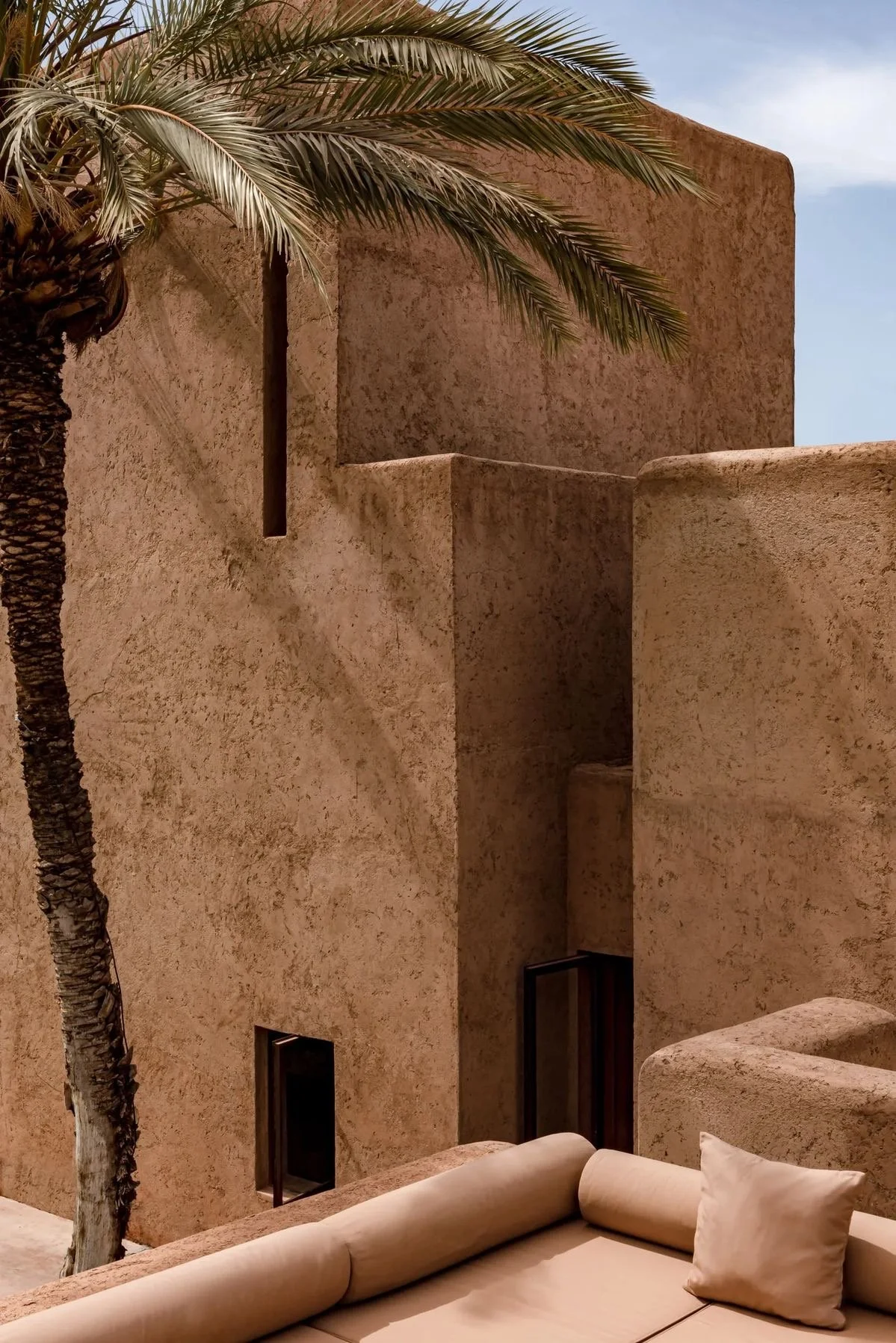 A palm tree beside a modern, beige stucco building with simple, clean lines and small windows, under a blue sky.