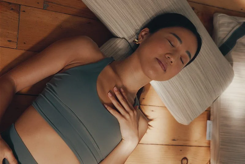 A woman lying on the floor with her head on a pillow, hand on chest, and eyes closed. 