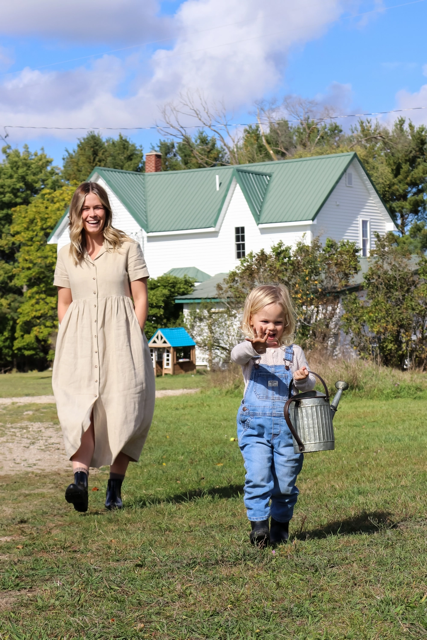 A blonde woman in a dress with boots walking behind a blonde child in denim overalls carrying a watering can outside