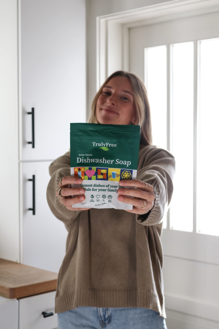 A blonde woman in a beige sweater showing the camera Truly Free Home Dishwasher Soap