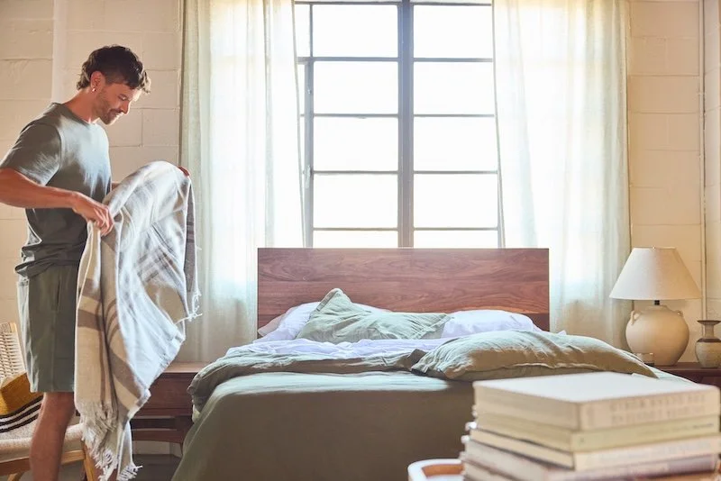 A standing man in a bedroom, holding a blanket to add to his bed.