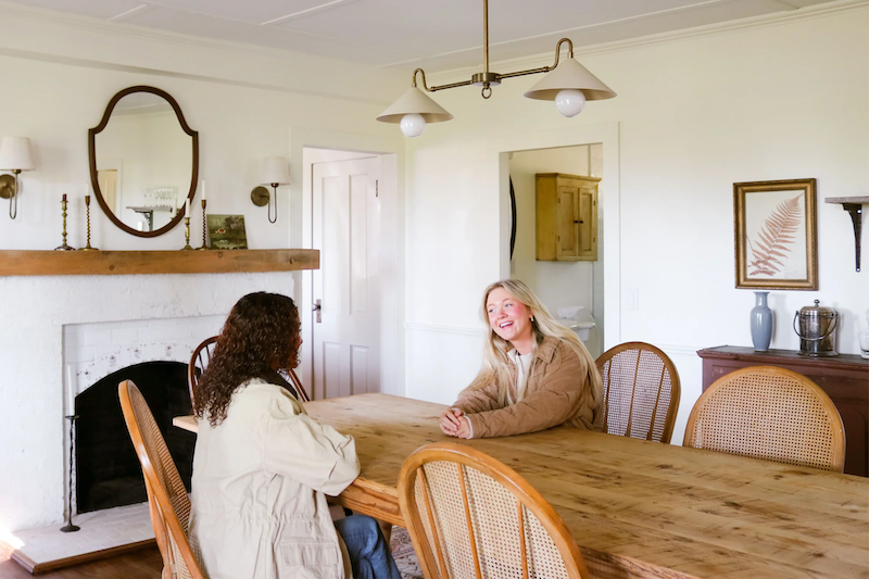 Two women wearing fall jackets sitting across from each other at a large wooden dining table