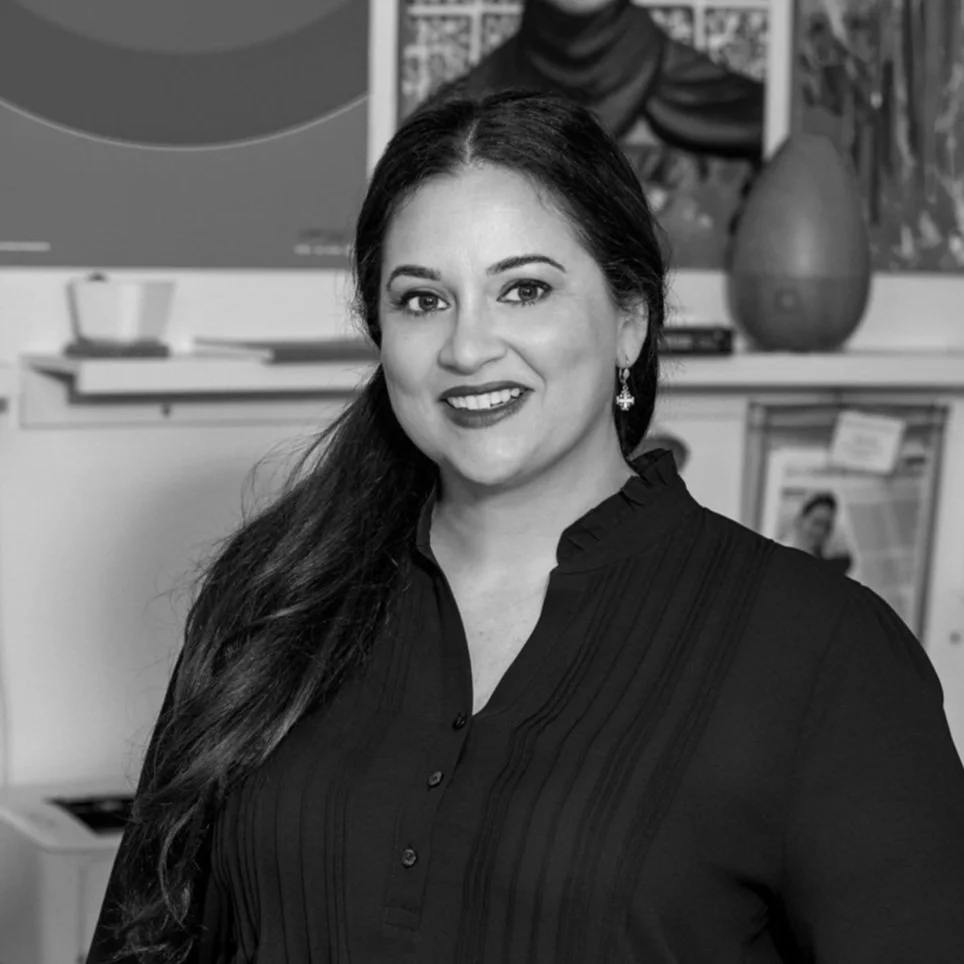 Black and white photo of a woman smiling in an office setting, with a bookshelf and various decorations in the background.