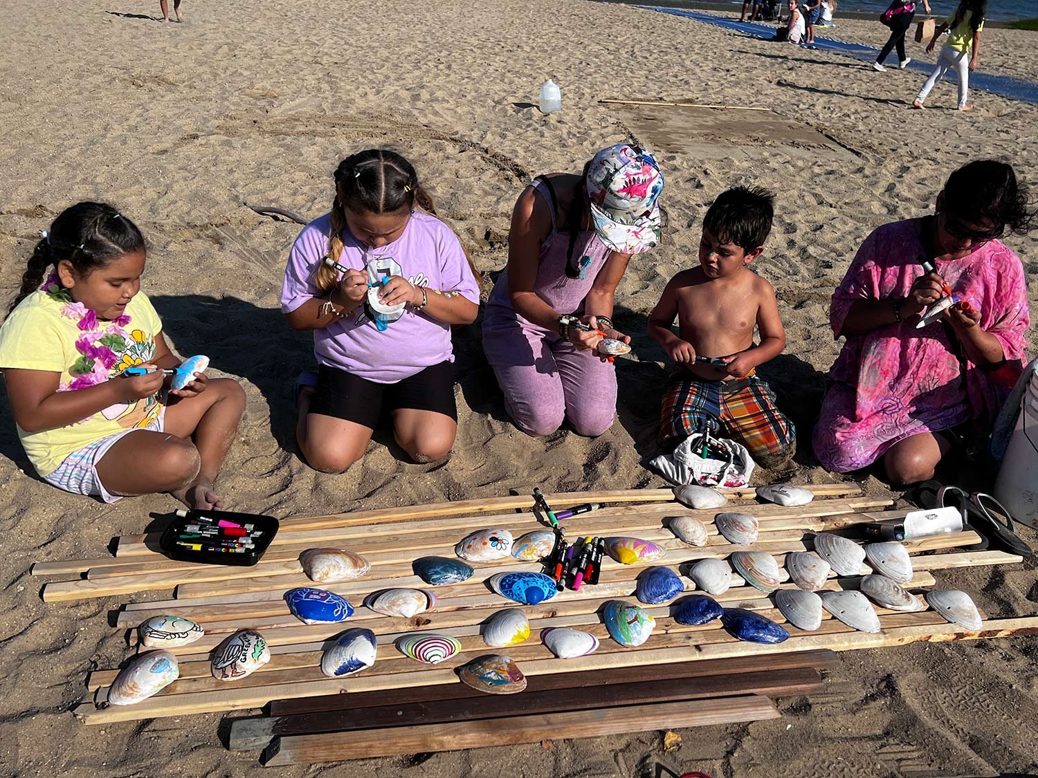 A group of children and adults sitting on sand decorating seashells with markers. There is a collection of painted shells laid out on wooden boards in front of them on the beach.