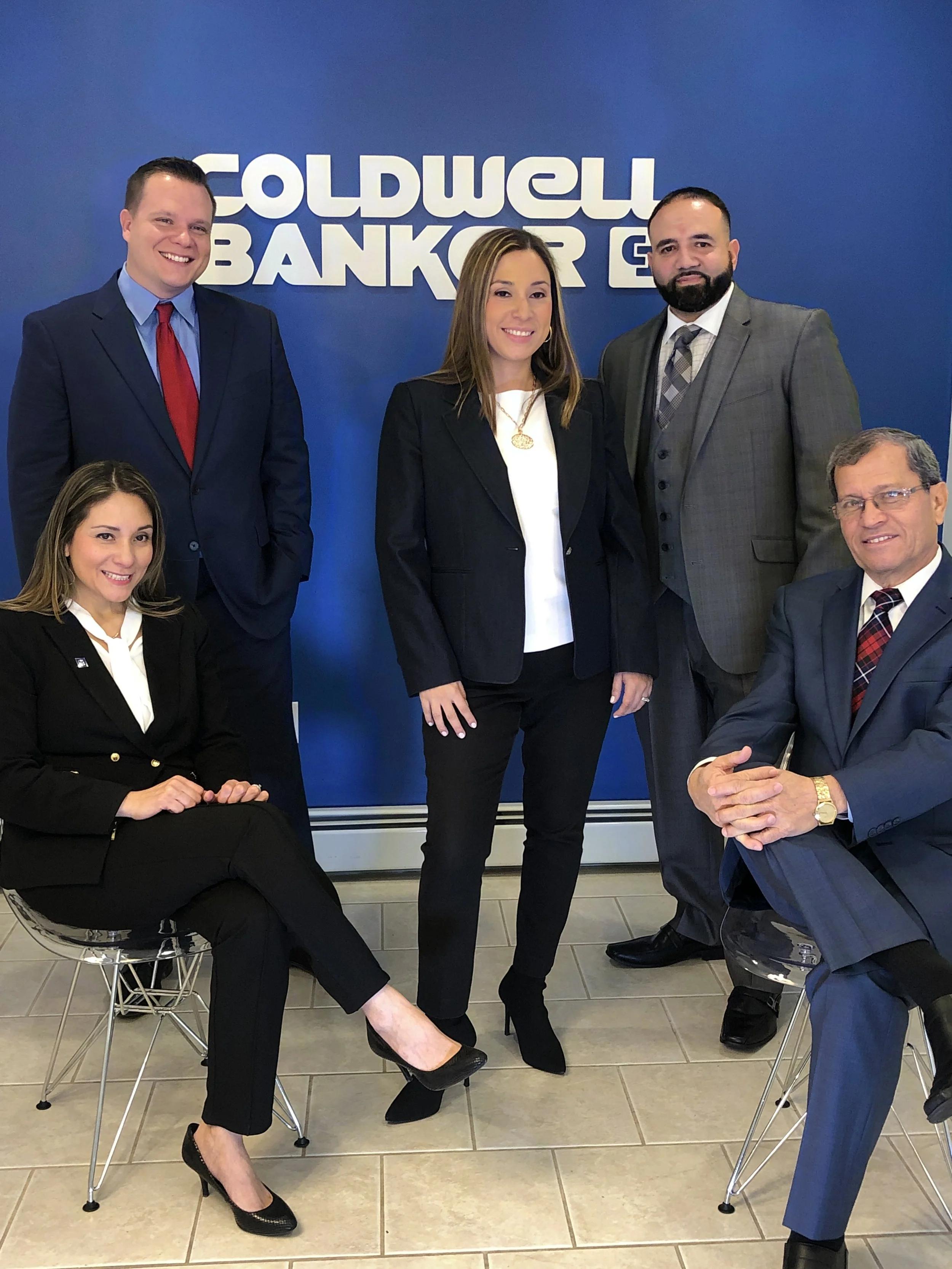 A group of five professionally dressed individuals standing and sitting in front of a Coldwell Banker sign, posing for a photo.