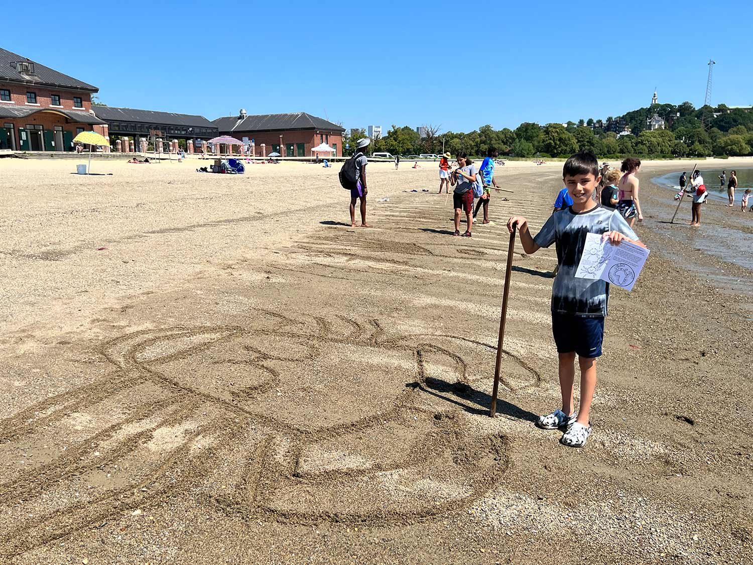 Boy holding a stick and paper standing on sandy beach with sea drawing, people and buildings in background.