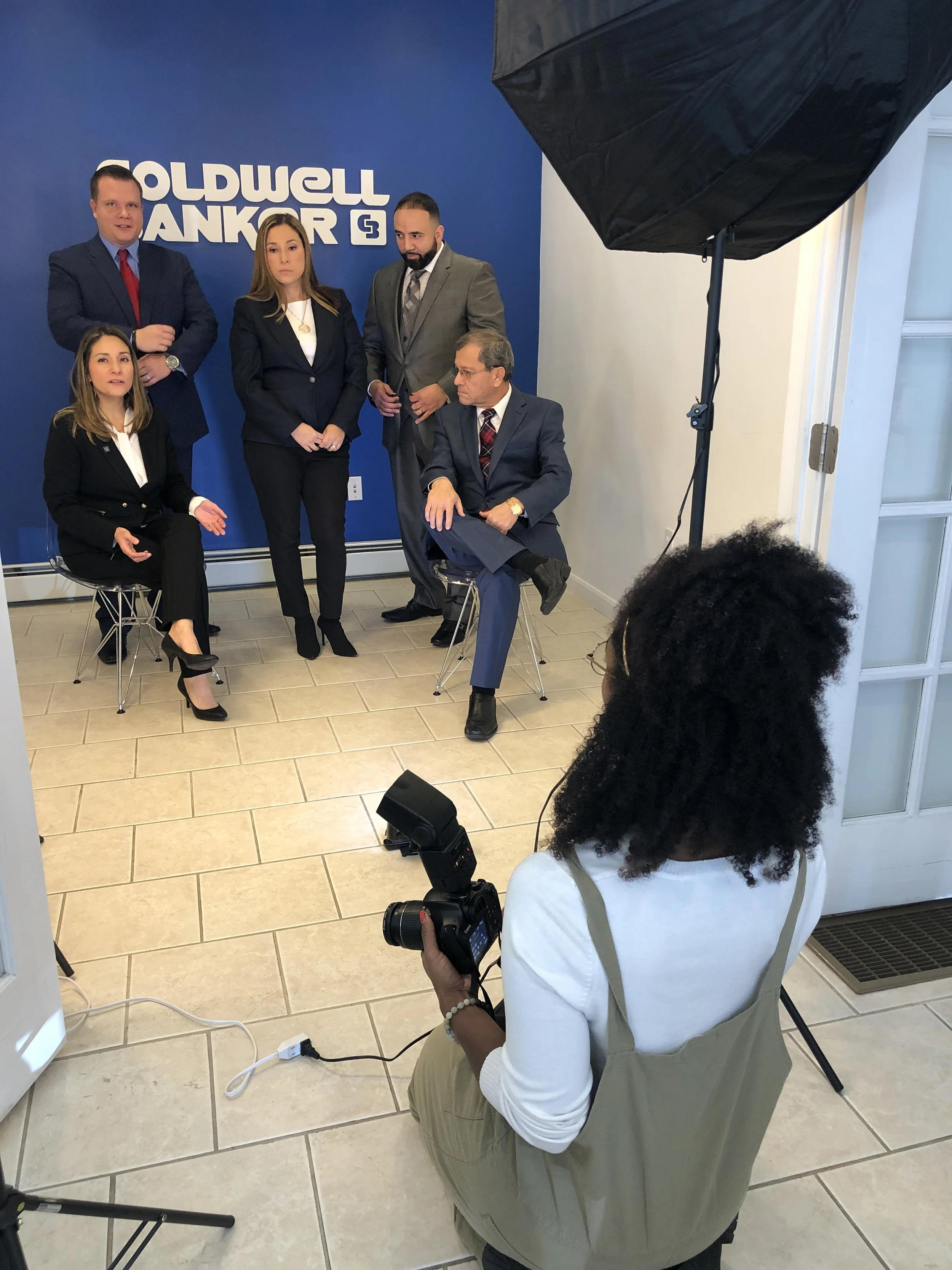 Group photo shoot in a professional setting with a photographer and five people in business attire, posing in front of a branded wall.