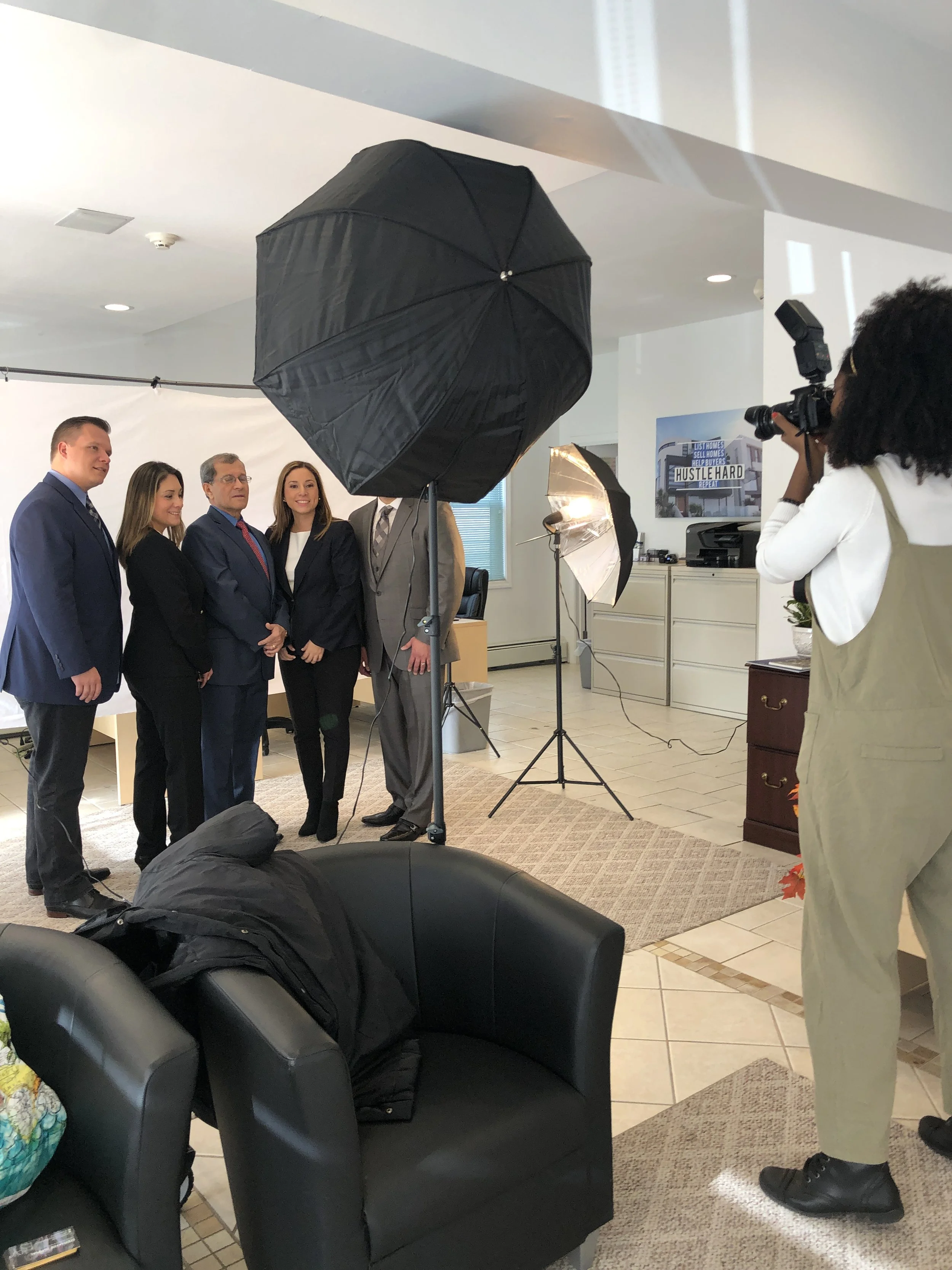 A group of business professionals posing for a photograph in an office setting, with a photographer capturing the scene. Studio lighting equipment and a backdrop are visible, along with office furniture.