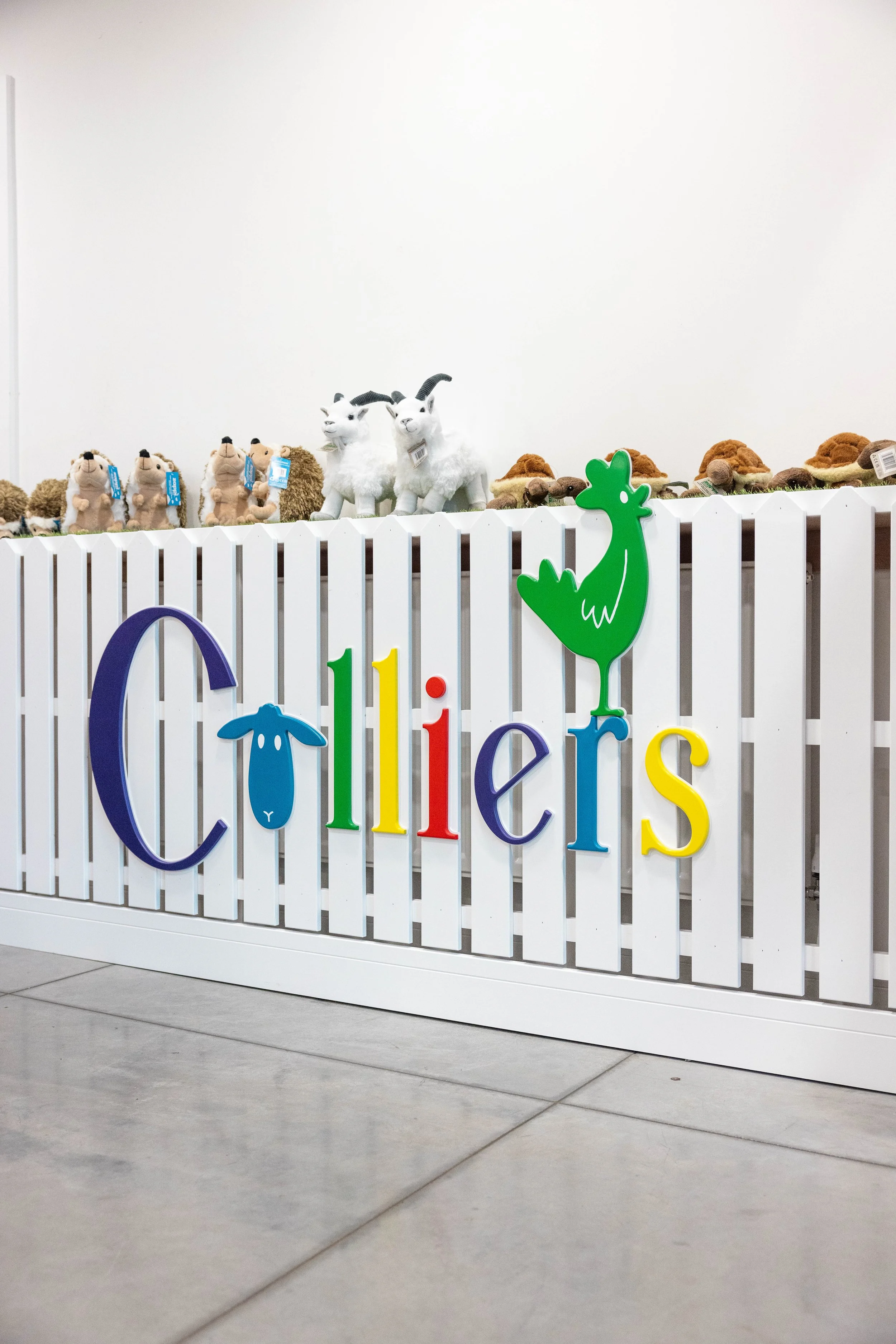 Display of stuffed animals with colorful letters spelling 'Chilliers' on a white picket fence, featuring a green dinosaur and a blue sheep.