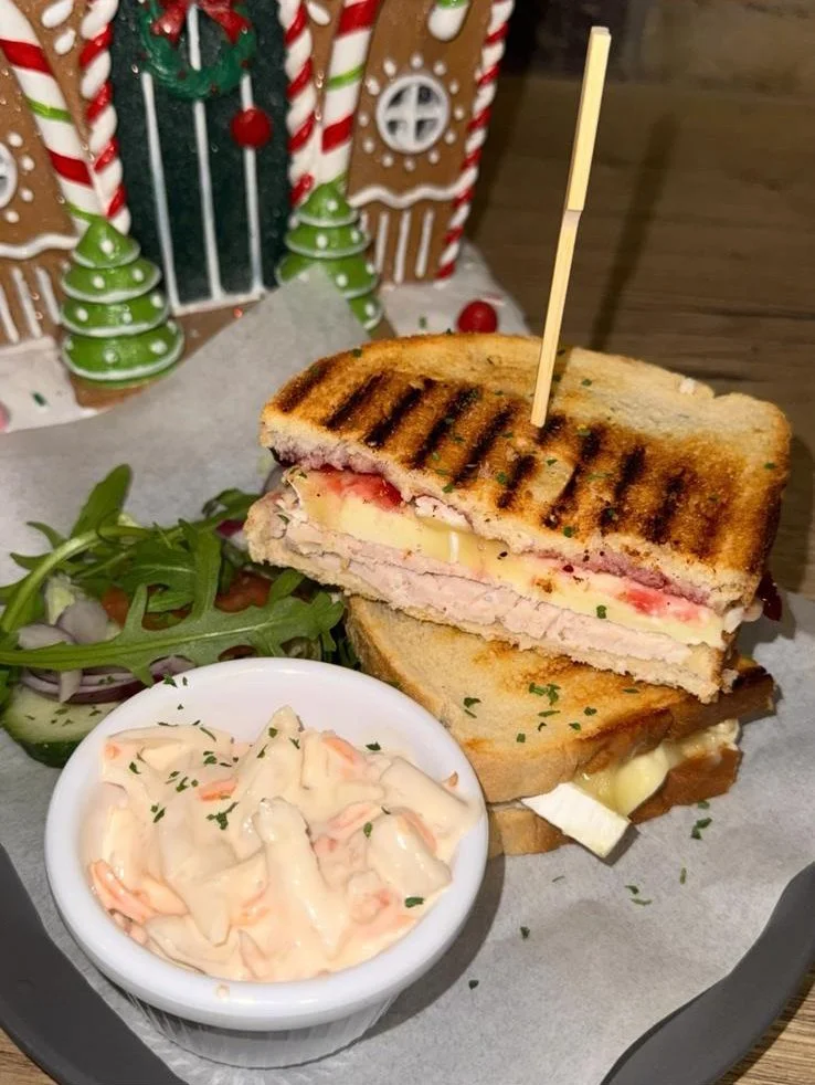 A grilled sandwich with turkey, cheese, and tomato, served with a side of coleslaw and a small salad, on a black tray with a Christmas-themed background.