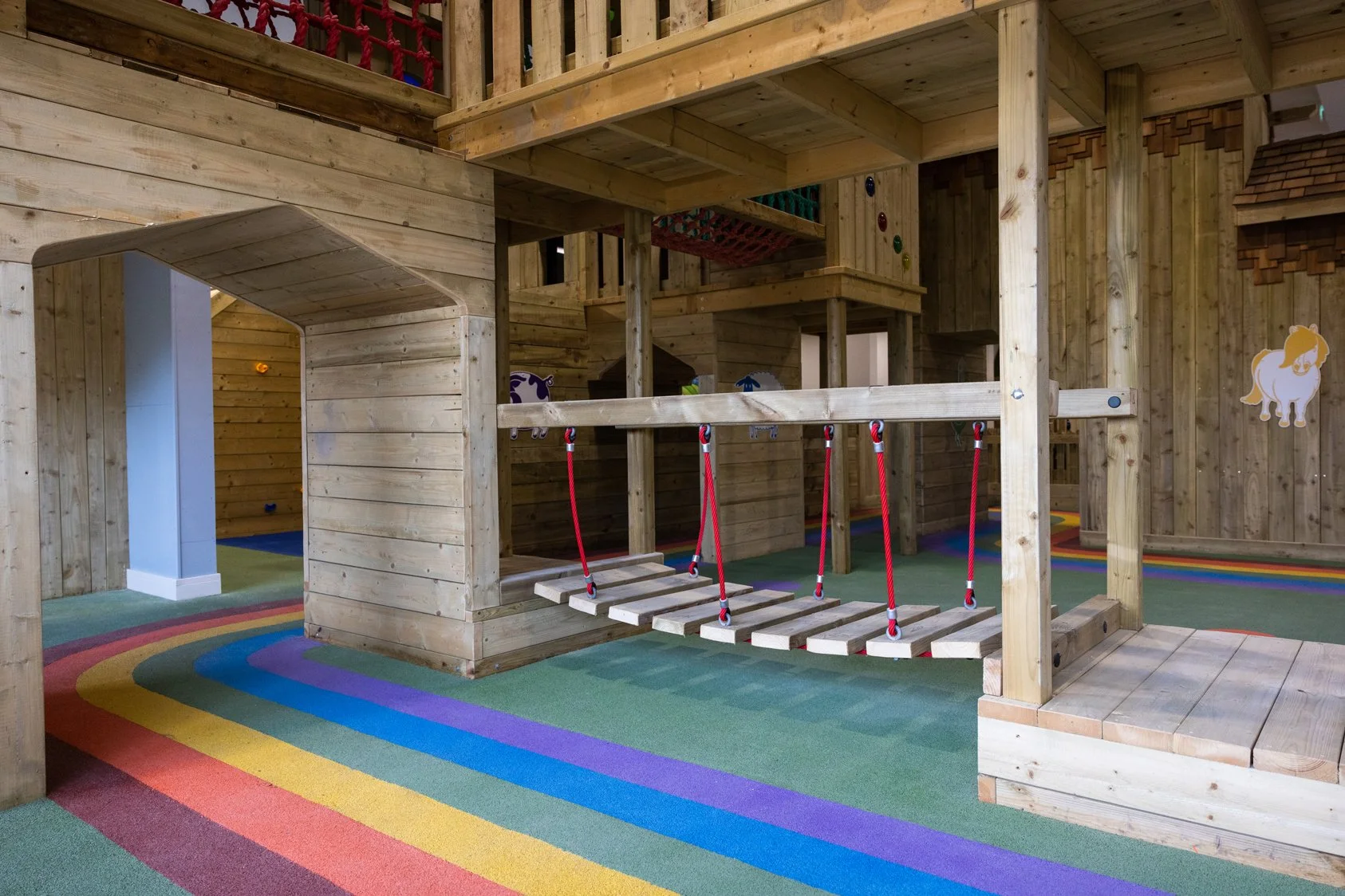 Indoor playground with wooden structures including a bridge with red ropes, colorful rainbow-striped carpet, and cartoon horse decals on the wooden walls.