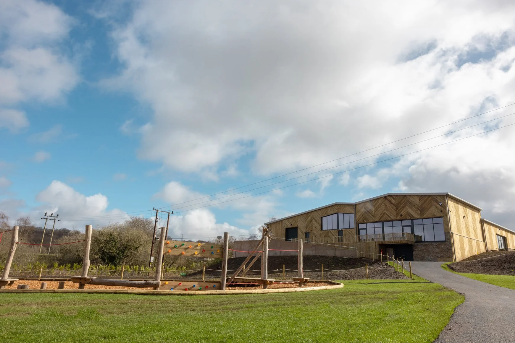 A modern building with wooden exterior and large windows, a playground with a wooden climbing structure and colorful climbing holds, a grassy lawn, a paved pathway, and a partly cloudy sky.