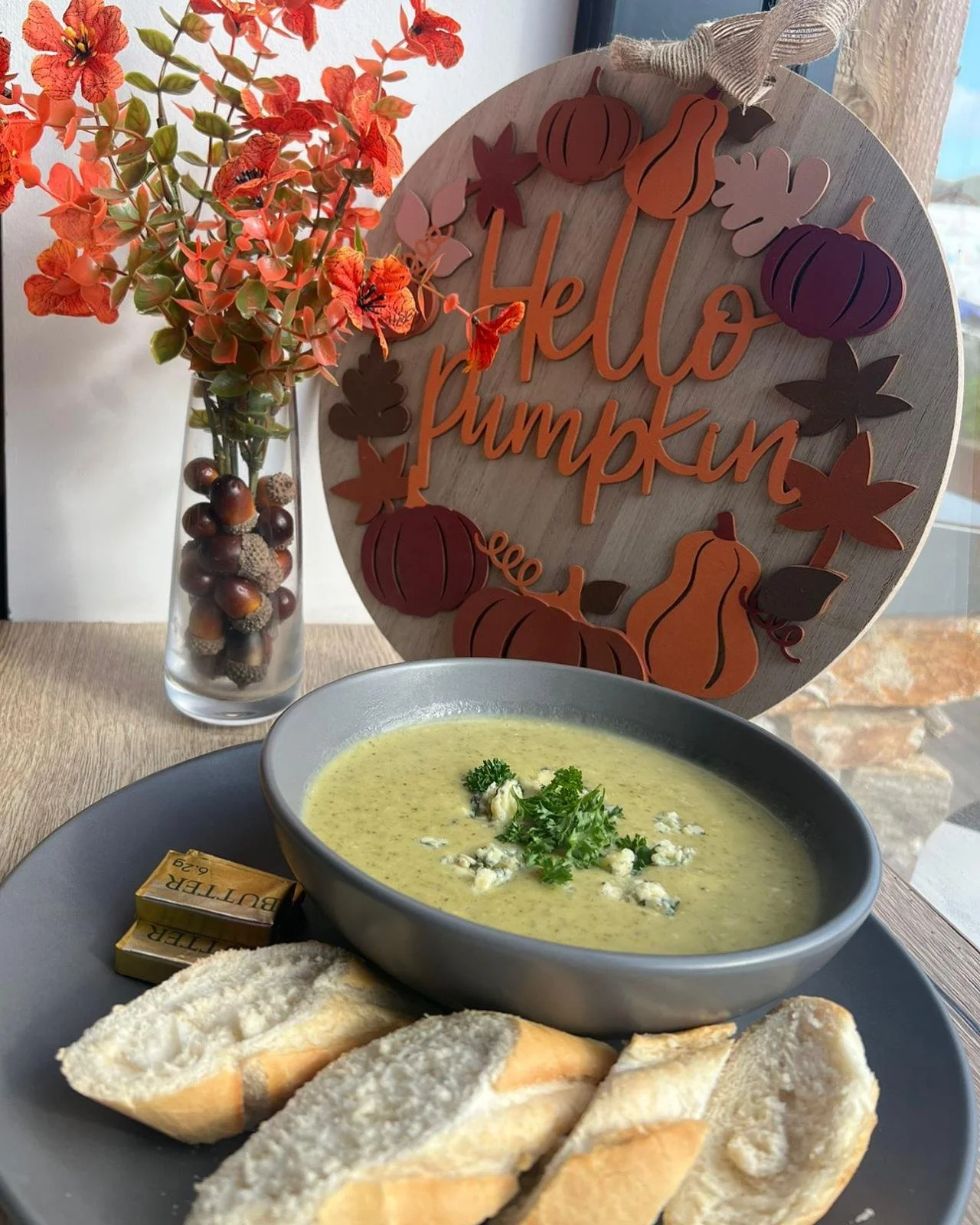 A bowl of greenish creamy soup garnished with herbs and cheese, served with sliced bread on a gray plate. Next to the plate are small butter packets. In the background, a flower vase with orange and pink flowers and a wooden sign with pumpkins and fall leaves that reads 'Hello Pumpkin' are visible.
