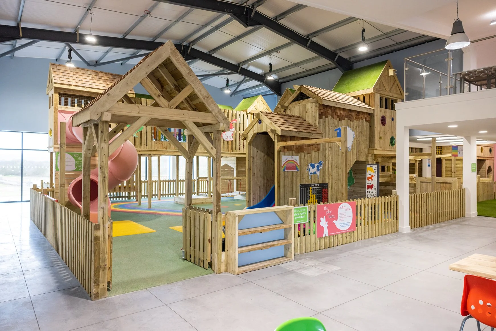 Indoor playground with wooden play structures, slides, and colorful decorations, enclosed by a low wooden fence.