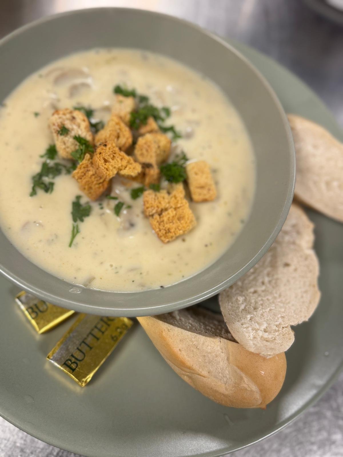 A bowl of clam chowder topped with croutons and parsley, served with oyster crackers and a small packet of butter on a gray plate.