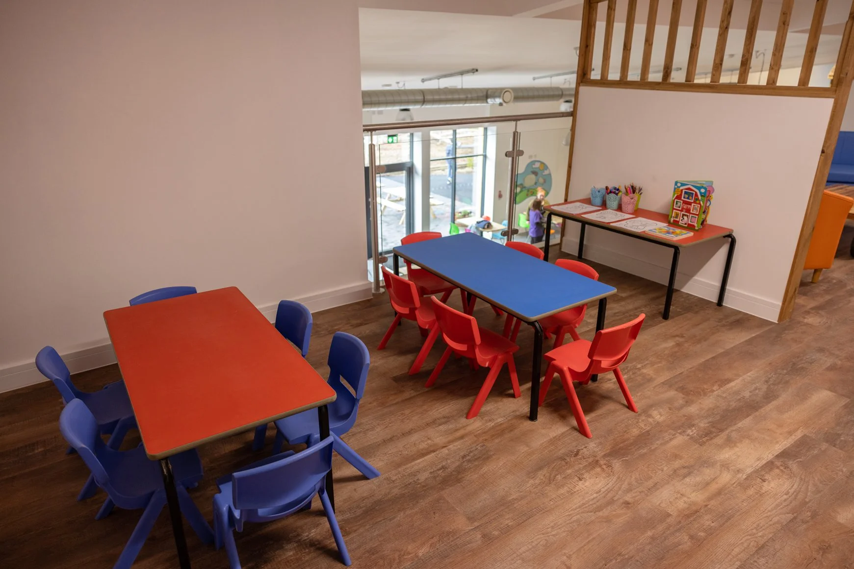 Children's play area with colorful tables and chairs, including a red table with blue chairs and a blue table with red chairs, near a window and a wall with a partial wooden railing.