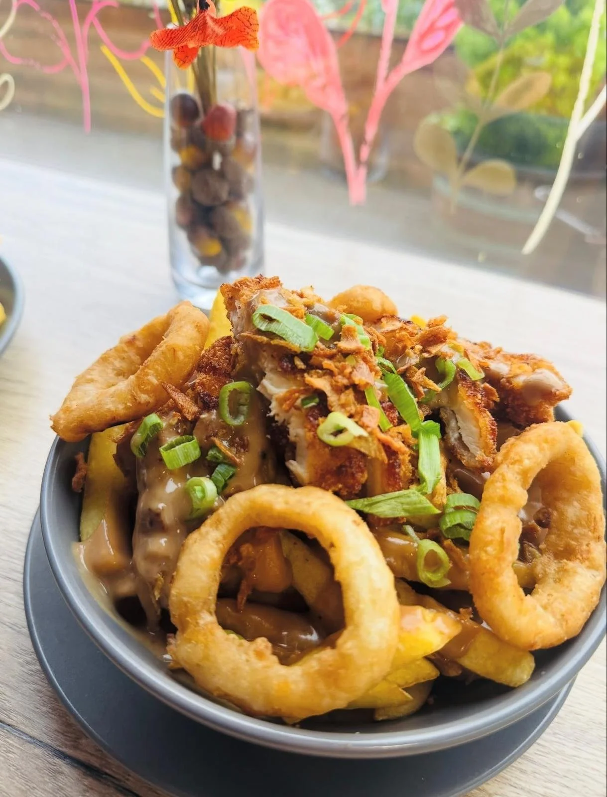 A bowl of poutine with French fries, cheese curds, brown gravy, fried chicken, green onions, and onion rings, on a wooden table with a floral window display in the background.
