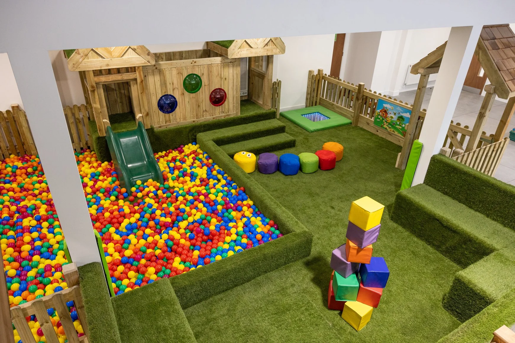 Indoor children's play area with a ball pit, slide, foam blocks, and soft grassy surfaces.