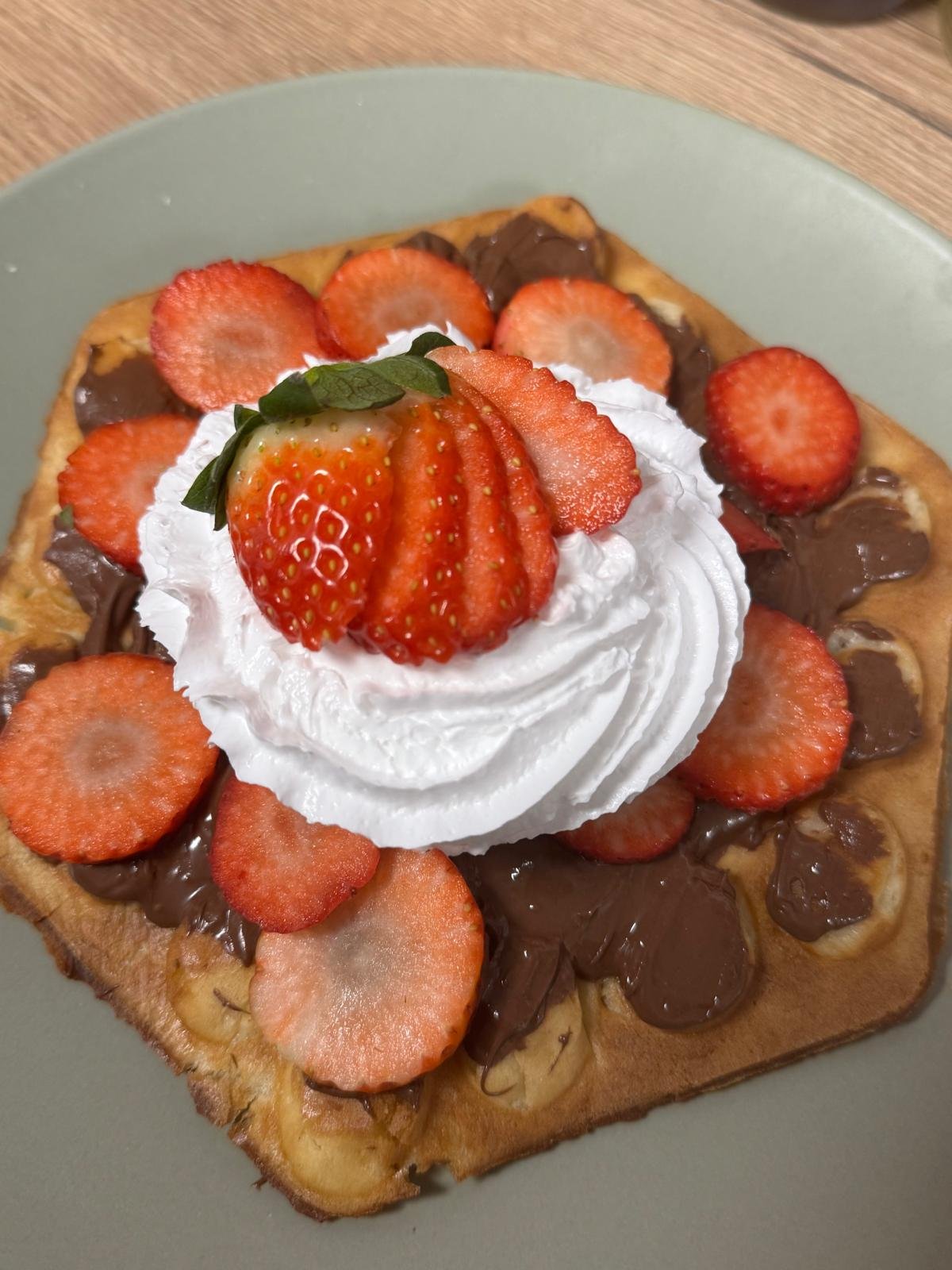 A square waffle topped with sliced strawberries, chocolate spread, whipped cream, and a whole strawberry with a mint leaf on top.