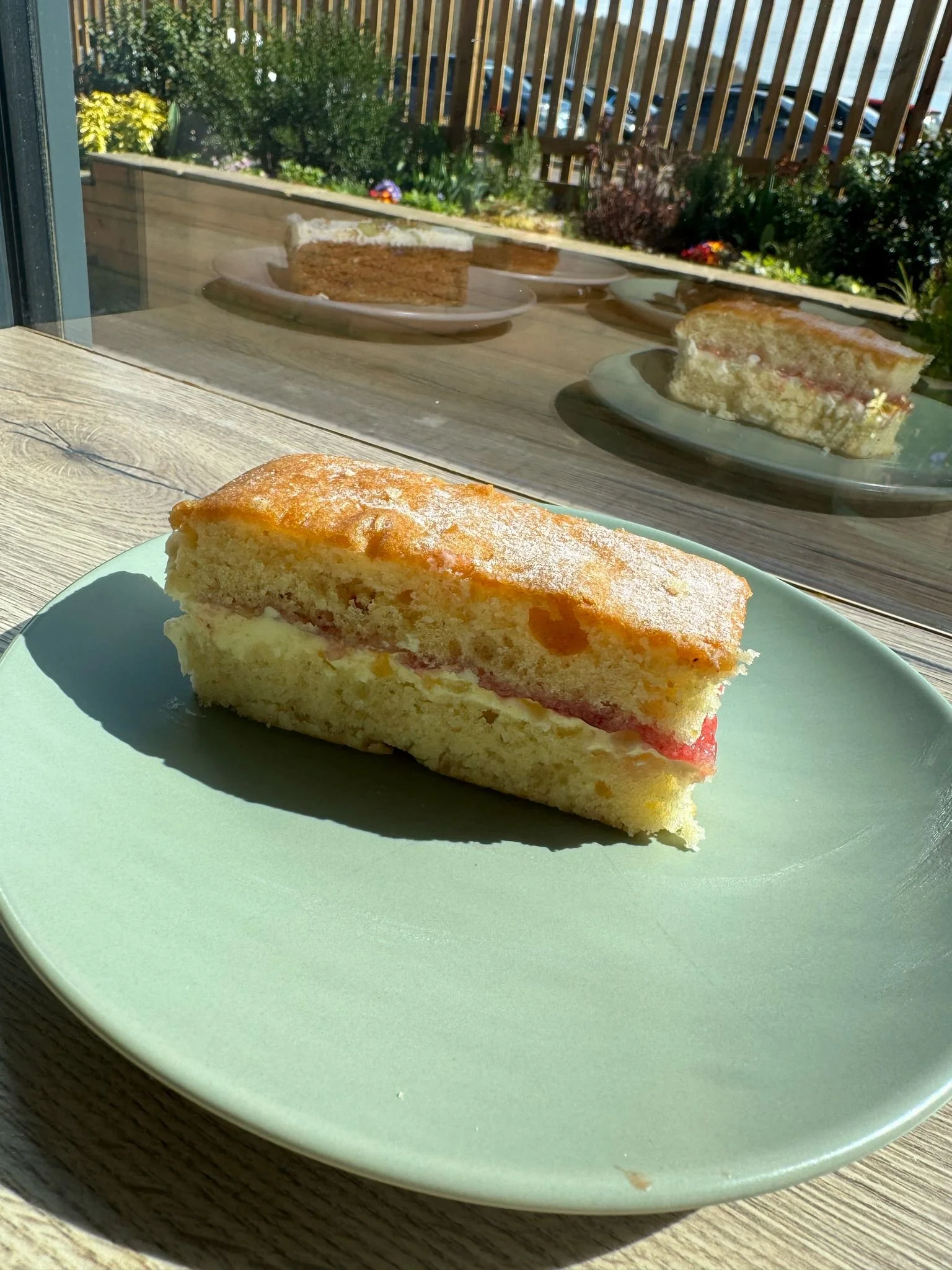 A slice of layered cake on a light green plate, with a window showing a garden outside. The cake has a yellow sponge, cream filling, and a pink or red fruit layer, with powdered sugar on top.
