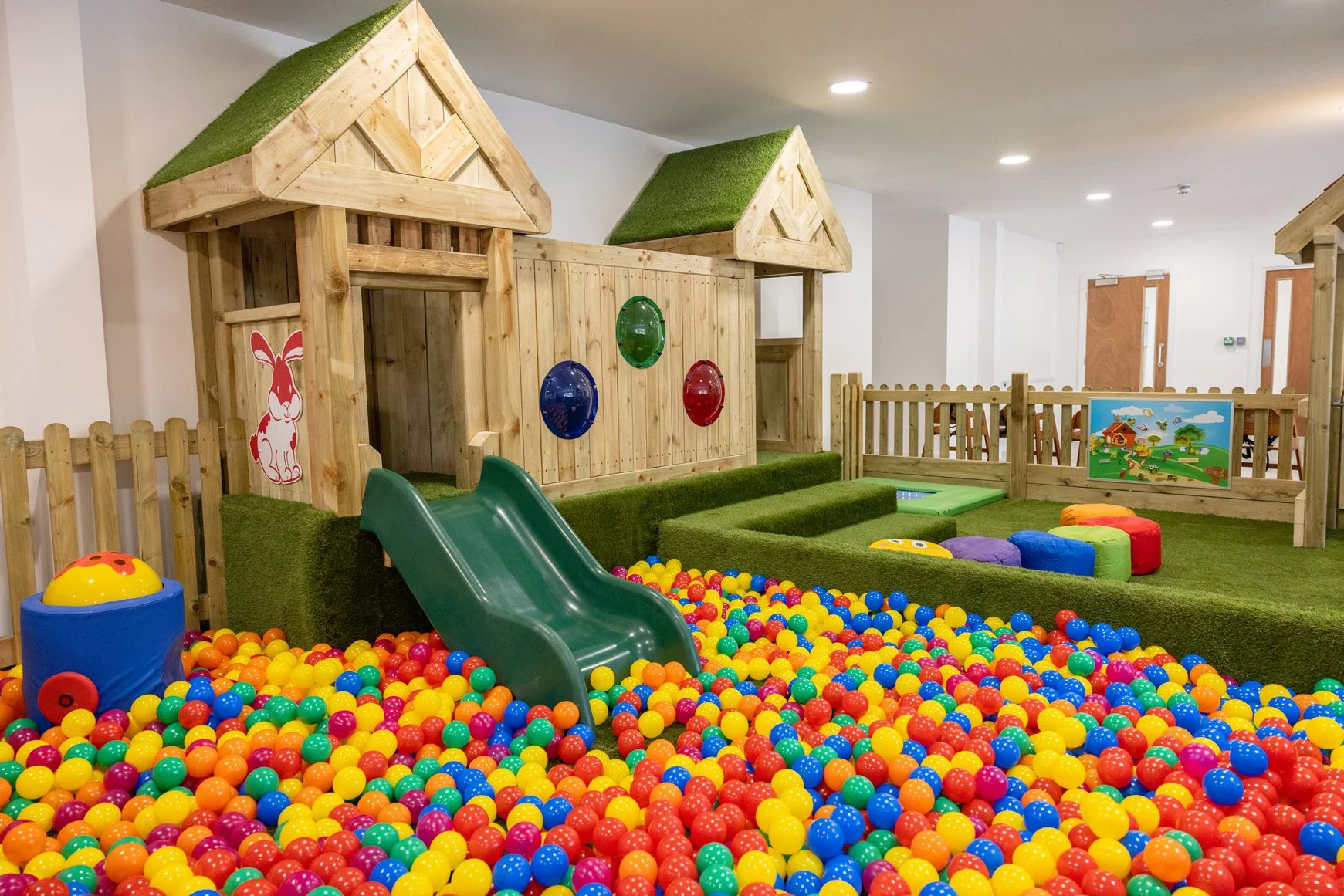 Indoor children's play area with a small green slide, colorful plastic balls, wooden play structures, and a fenced play zone with a colorful scene on a board.