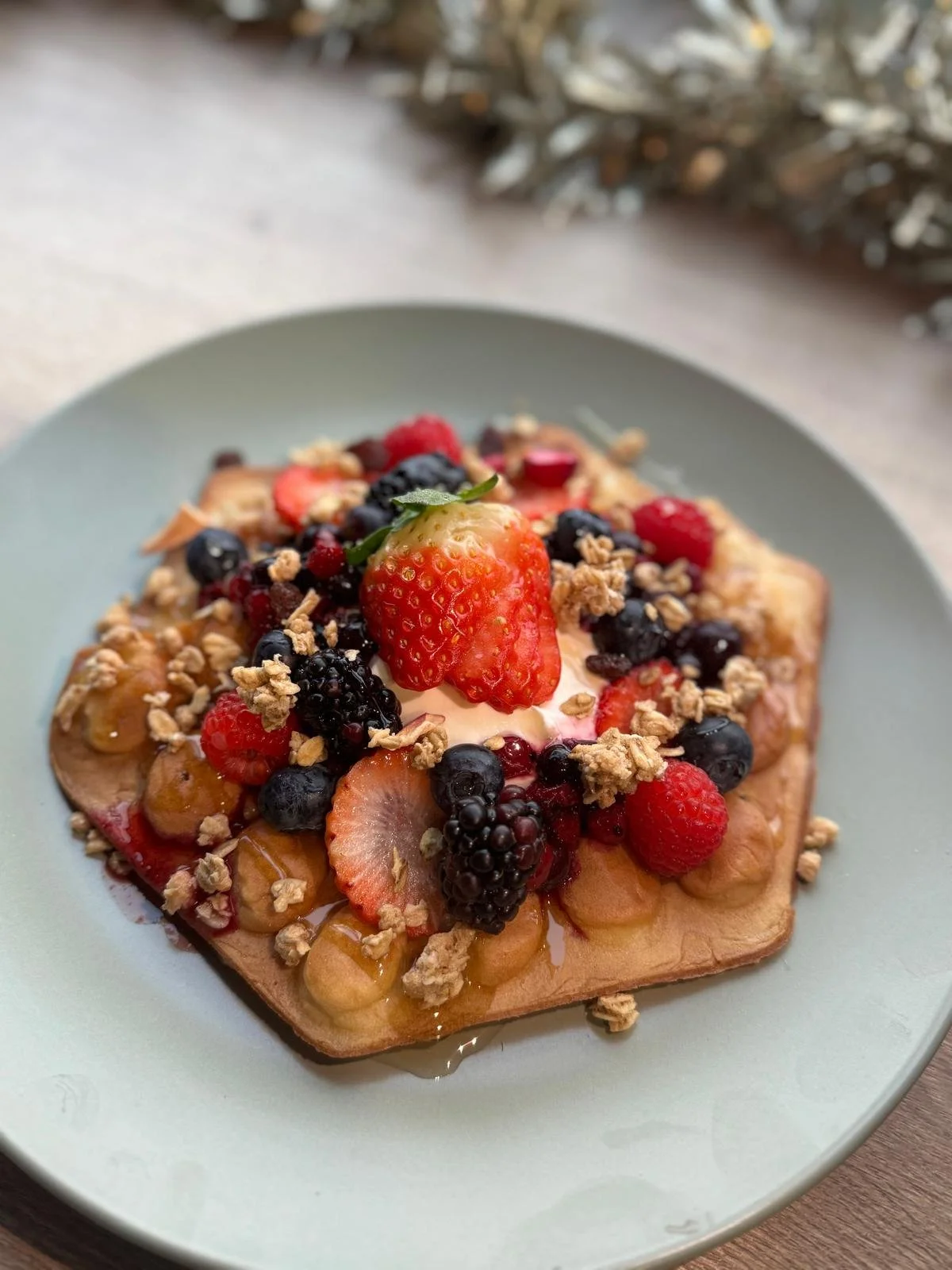 A waffle topped with various fresh berries, granola, and a dollop of cream or yogurt, served on a light blue plate.