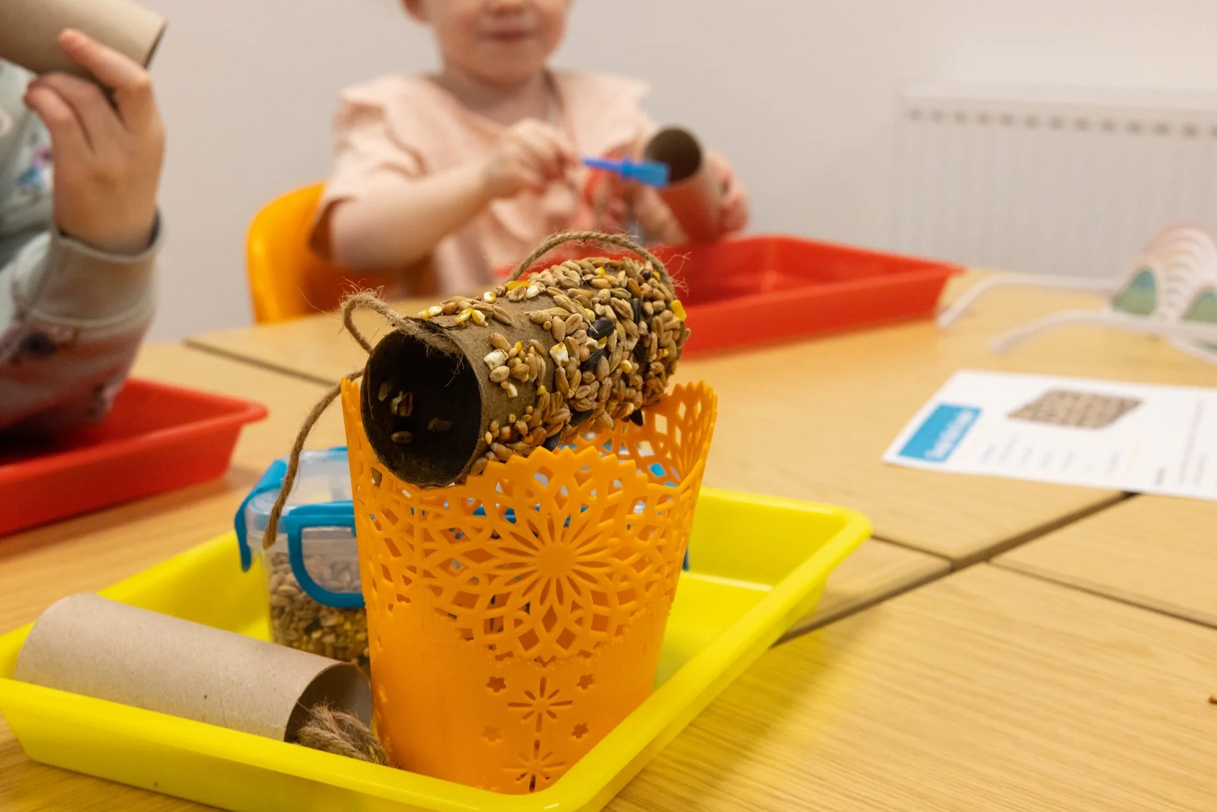 A craft project featuring a toilet paper roll covered in seeds, placed on an orange lantern with a yellow tray underneath, on a wooden table. A child in the background holds a similar craft, with other craft materials visible.