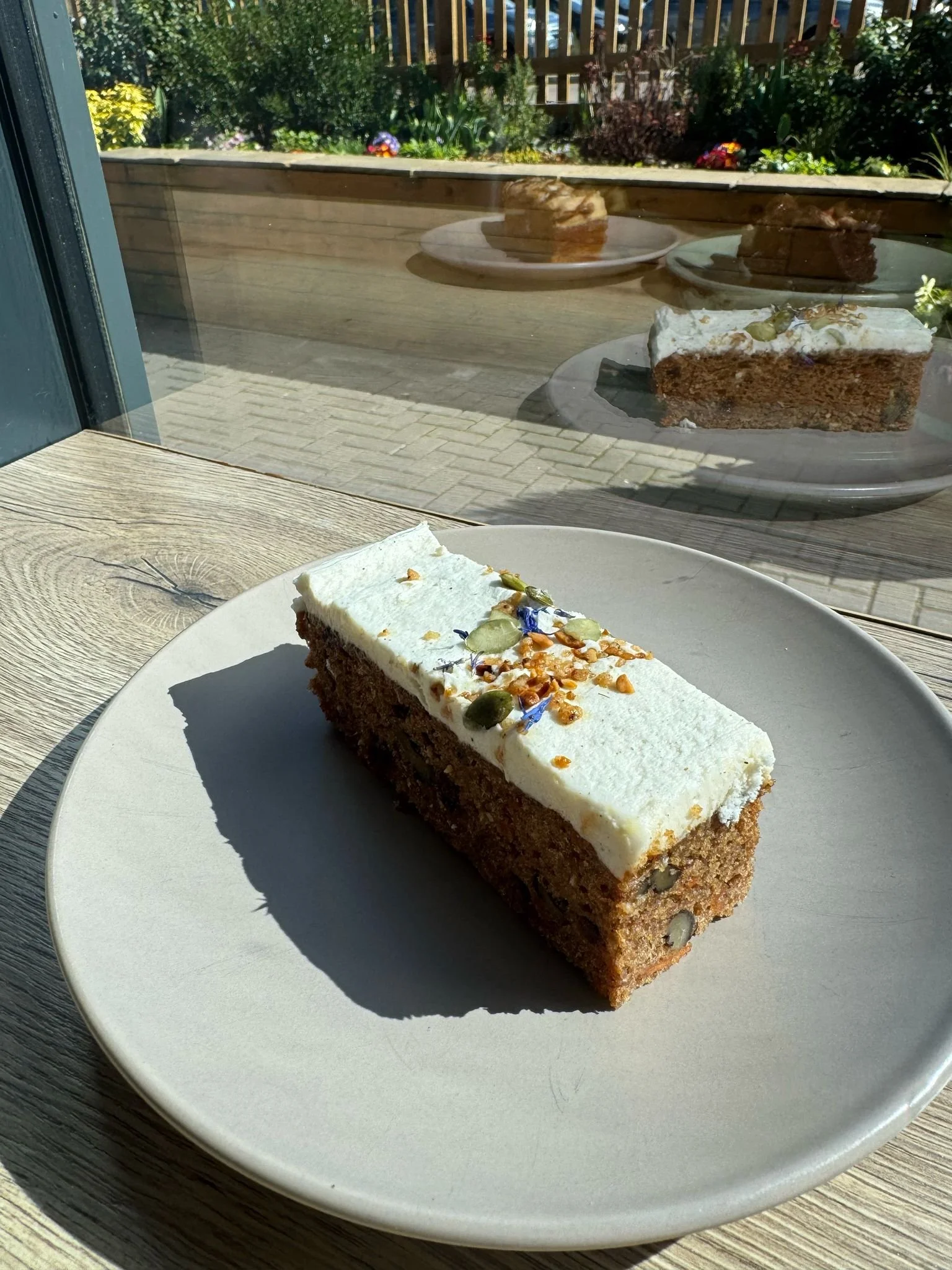 A slice of carrot cake with cream cheese frosting, topped with seeds and flower petals, on a white plate near a window, with more slices of cake on plates outside.