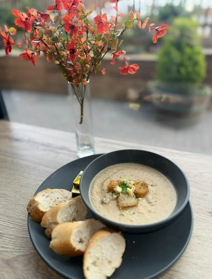 A black bowl of creamy mushroom soup garnished with croutons and chopped green onions, accompanied by sliced baguette bread on a black plate. In the background, a glass vase with pink and red artificial flowers is placed on a light wood table, with a window showing an outdoor scene.